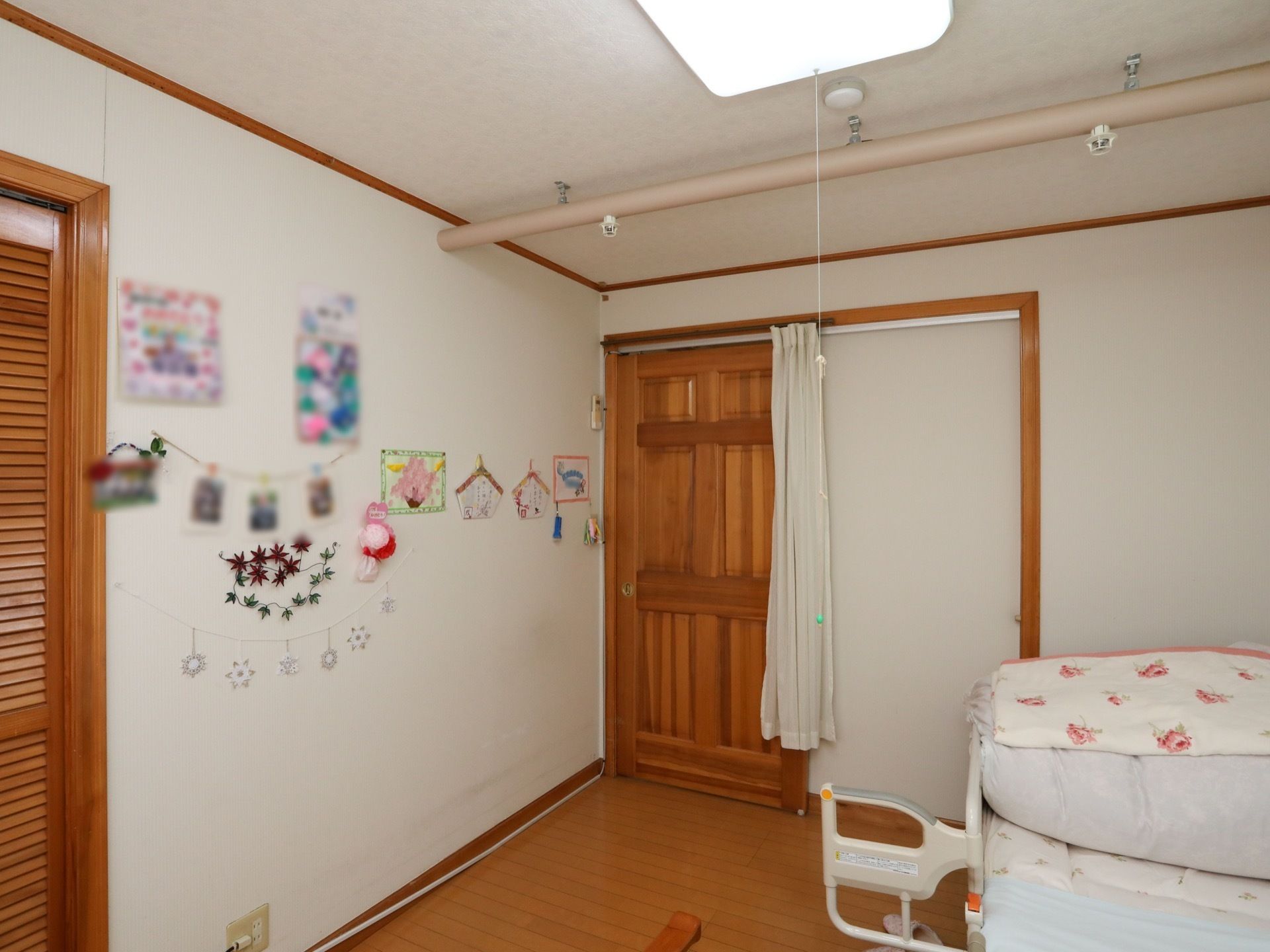 居室の床はフローリングで、柵が付いた介護用ベッドがある。白い壁にさまざまな作品が飾られている。
