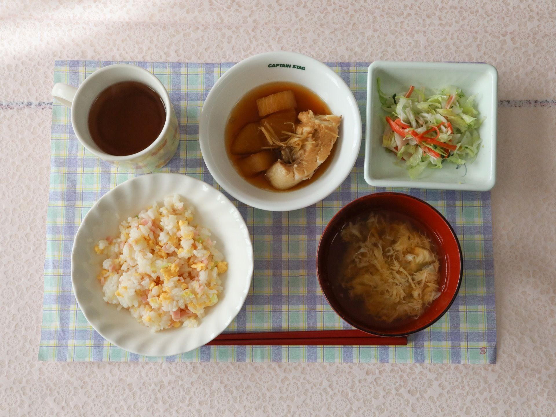 水色のチェックのランチョンマットに、ご飯、たまごスープ、魚の煮つけ、サラダ、お茶、箸が並べられている。