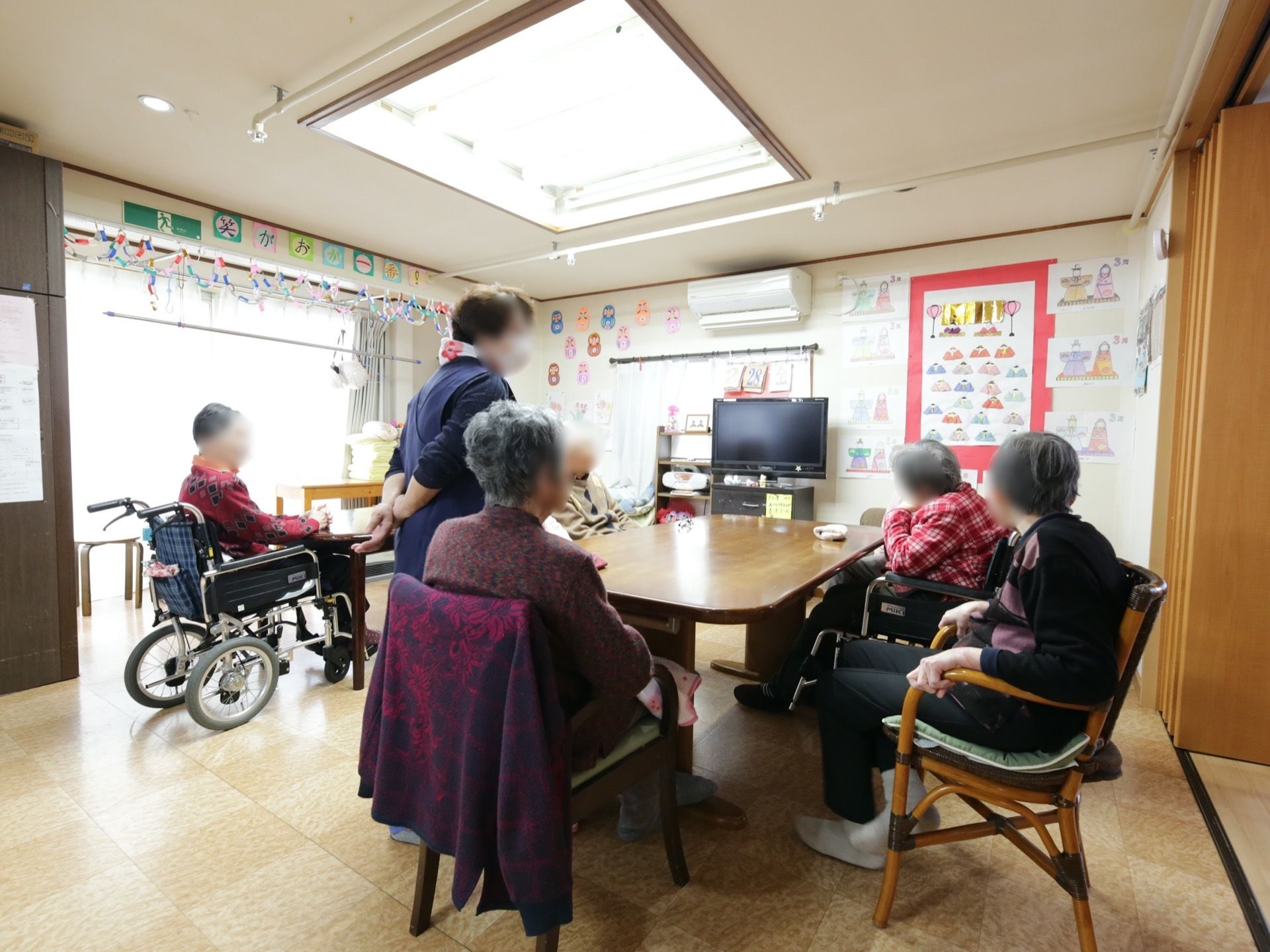 ダイニングテーブルが置かれたスペースがあり、入居者様が集まっている。壁際にはテレビが設置され、窓からは外光が入っている。
