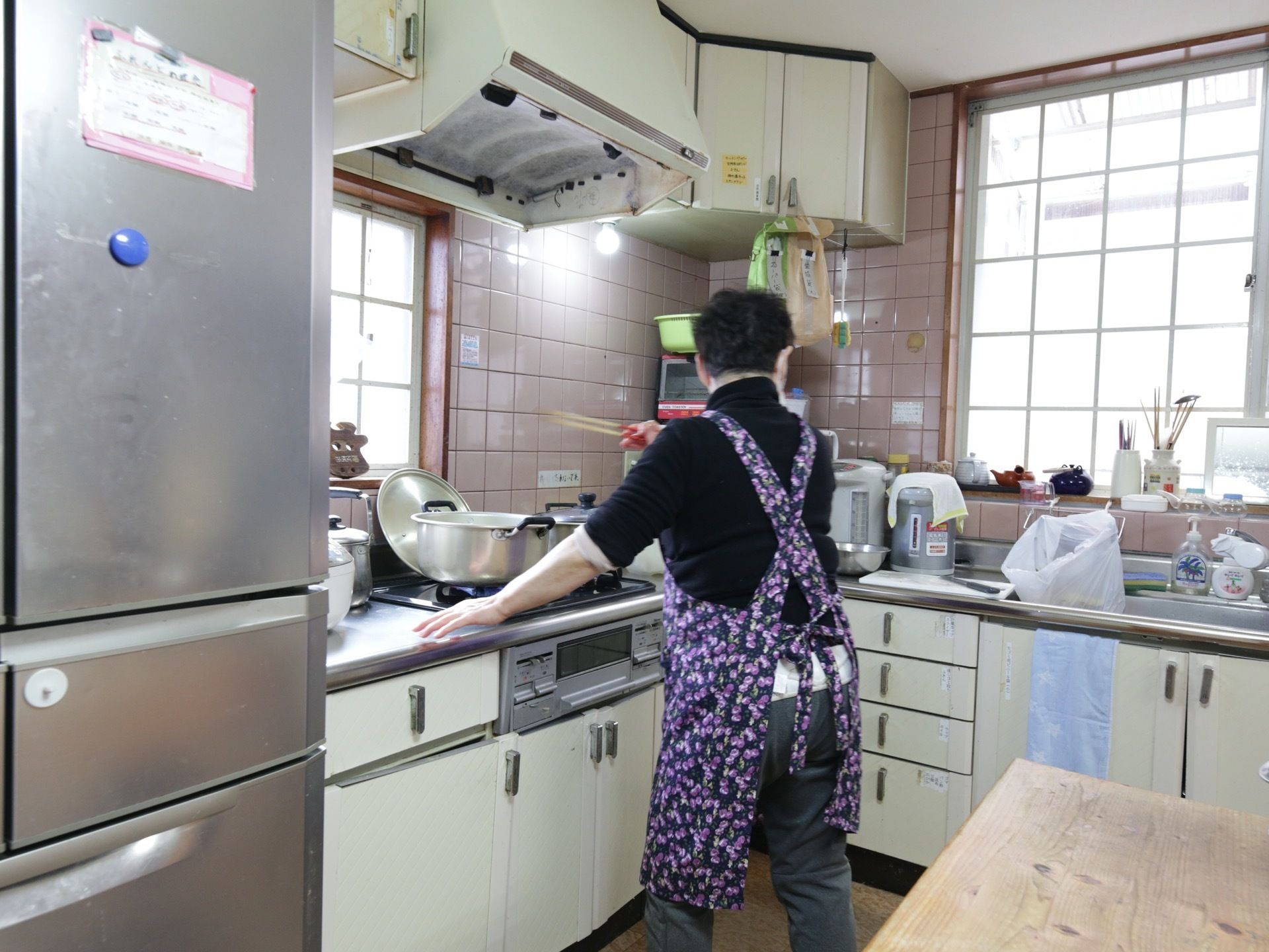 ガスコンロが２口付いたキッチンで、女性スタッフが調理を行っている。近くには作業台や冷蔵庫が設置されている。