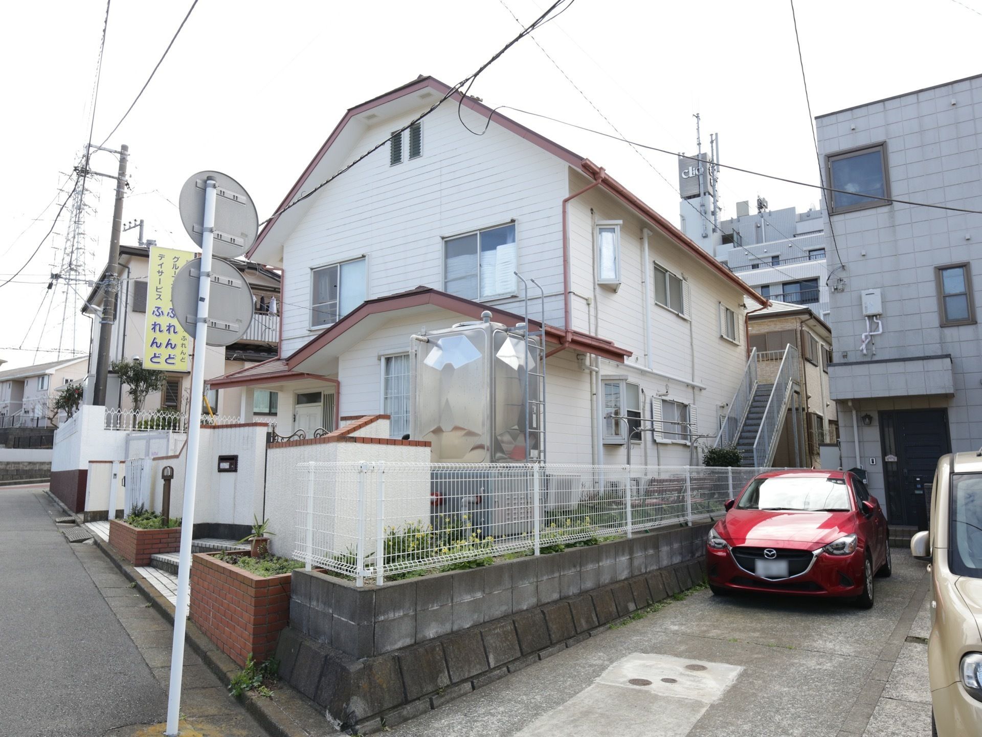住宅街の中に白い壁の戸建て住宅が建っている。敷地は白いフェンスで囲まれ、入り口には施設の看板が設置されている。