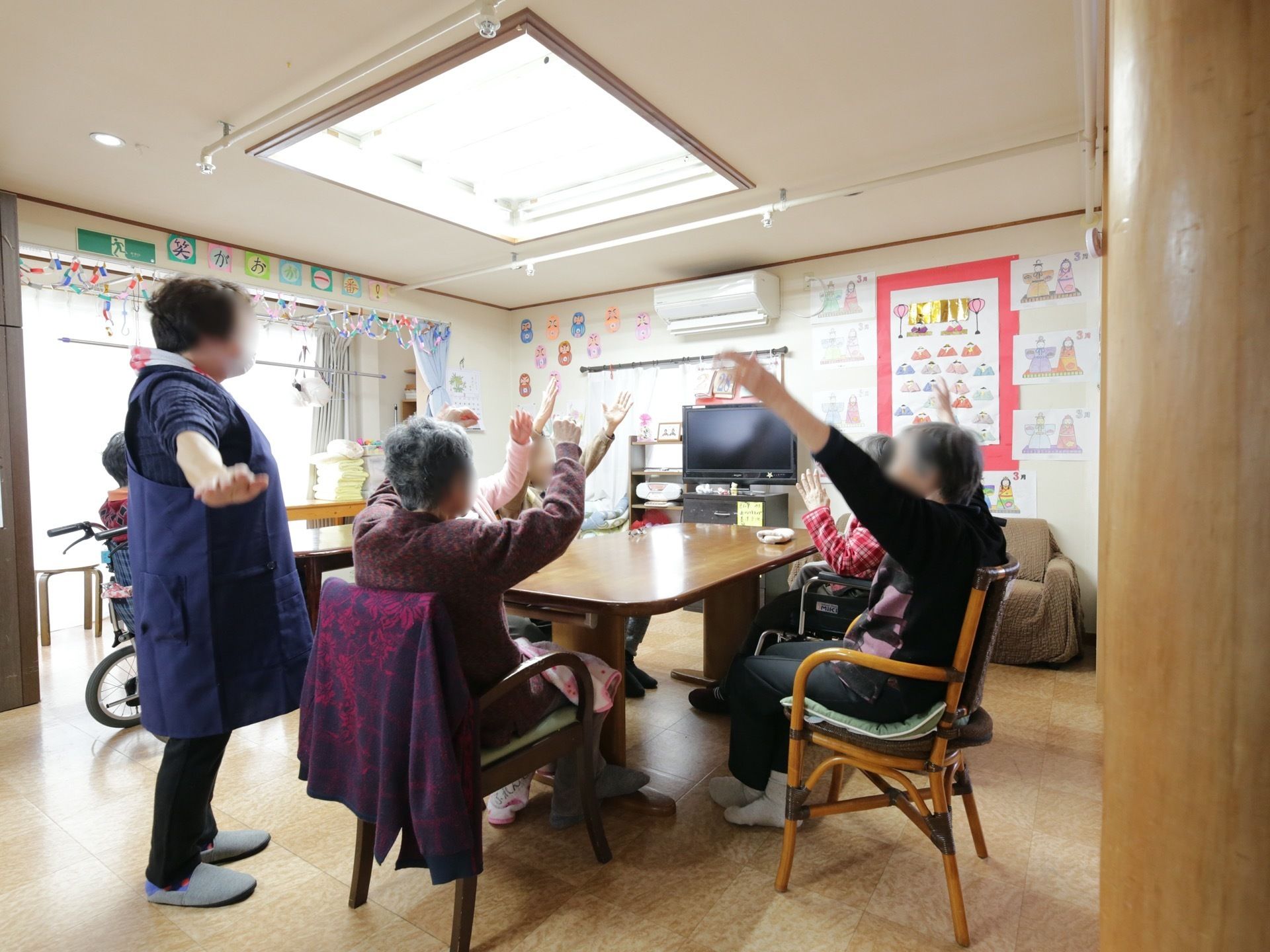 掃き出し窓のある明るいリビングに、テーブルセットが置かれている。入居者様が席について手を動かす運動を行っている。