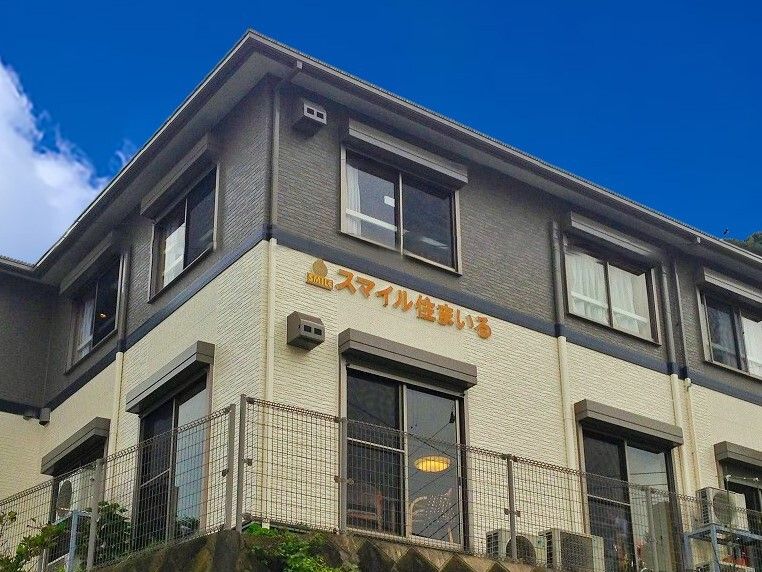 ダークグレーとベージュの外壁の2階建ての建物。建物の周りにはフェンスが建てられ、低木も植えられている。1階の大きな窓の向こうに共有スペースがある。