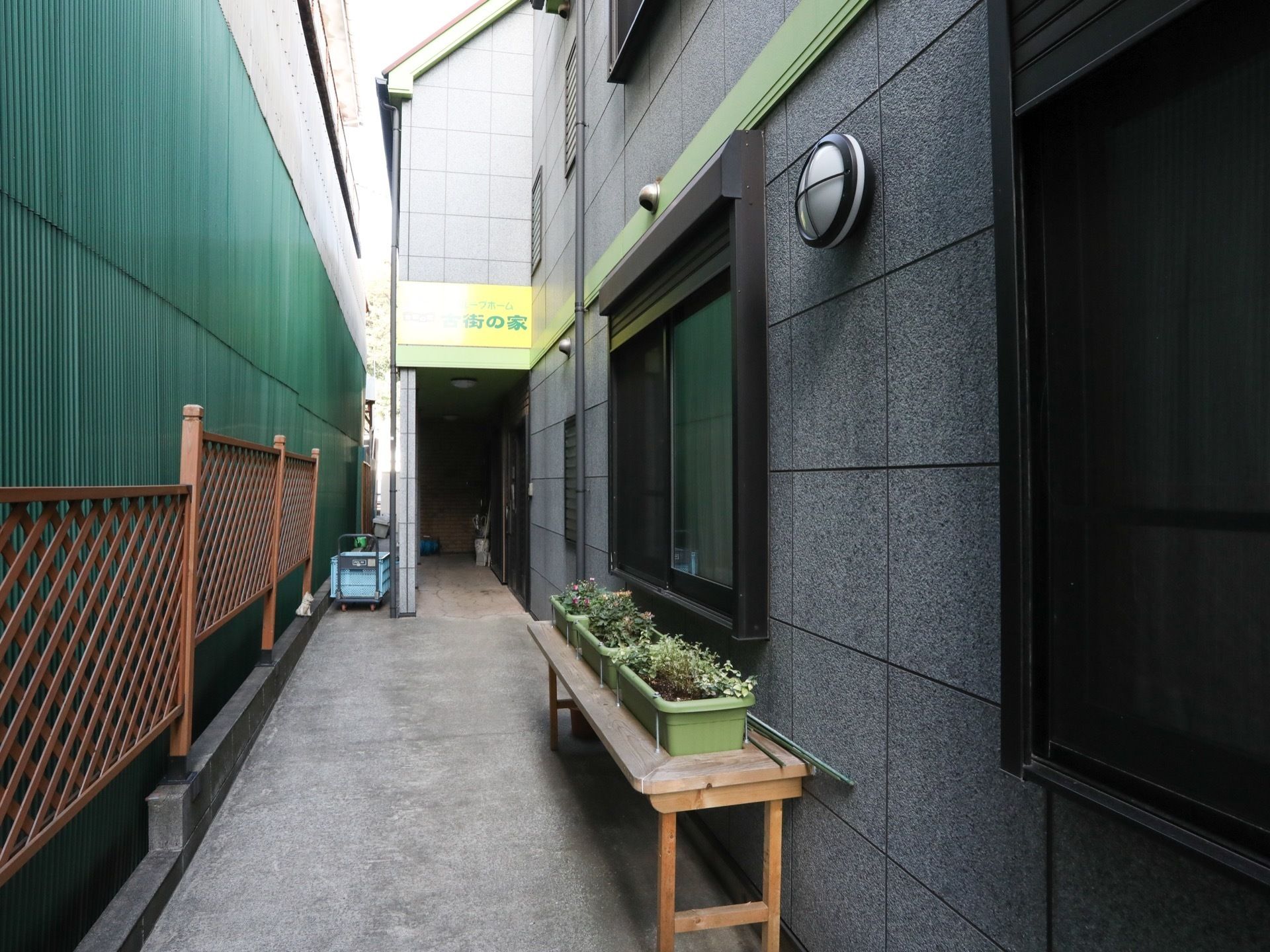 玄関へ通じる道はコンクリートが敷かれている。施設の横にベンチが置かれており、ベンチの上にプランターが飾られている。