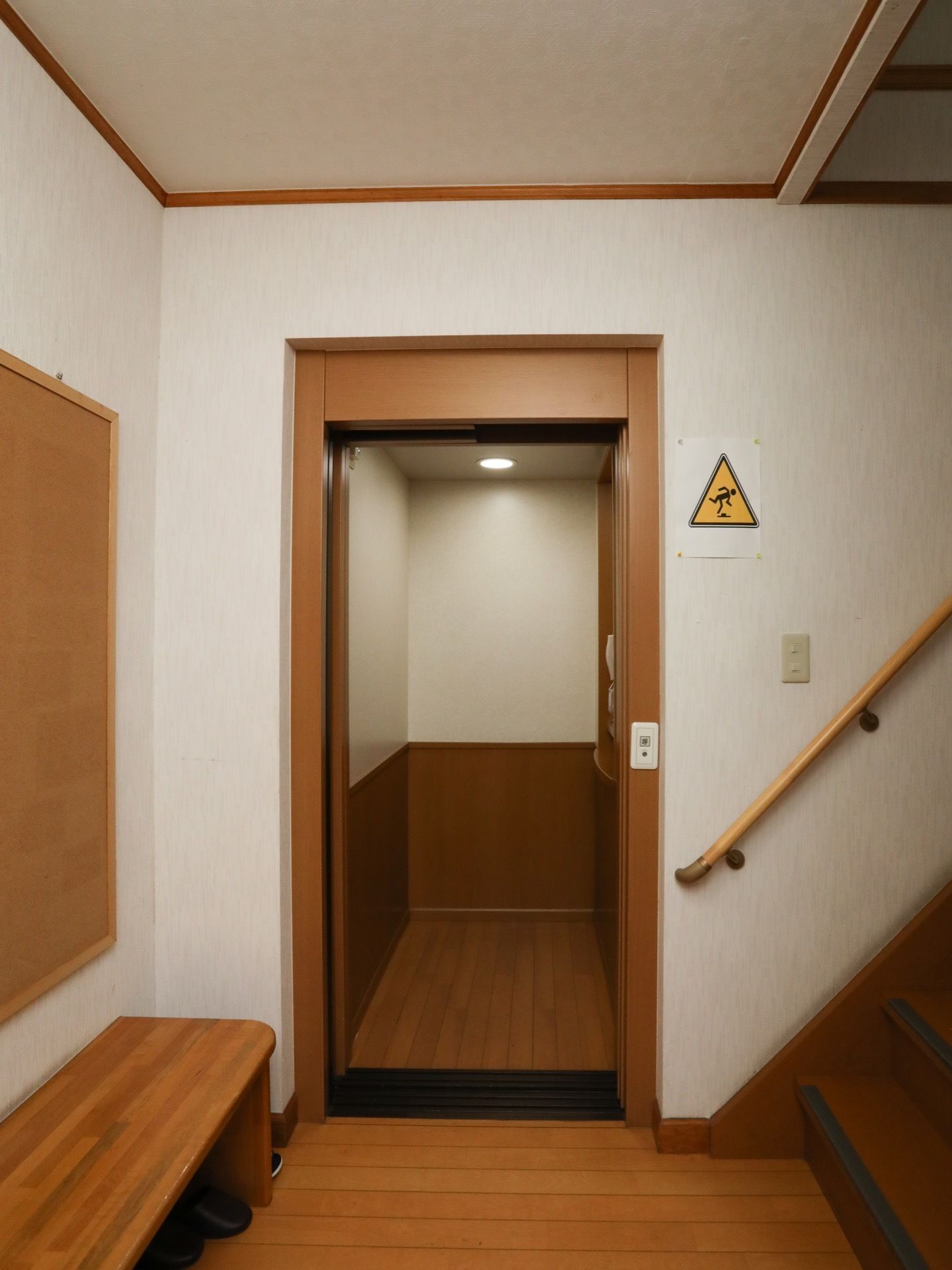 階段の前にエレベーターが設置されており、扉が開いている。エレベーターの前には、木目調のベンチが置かれている。