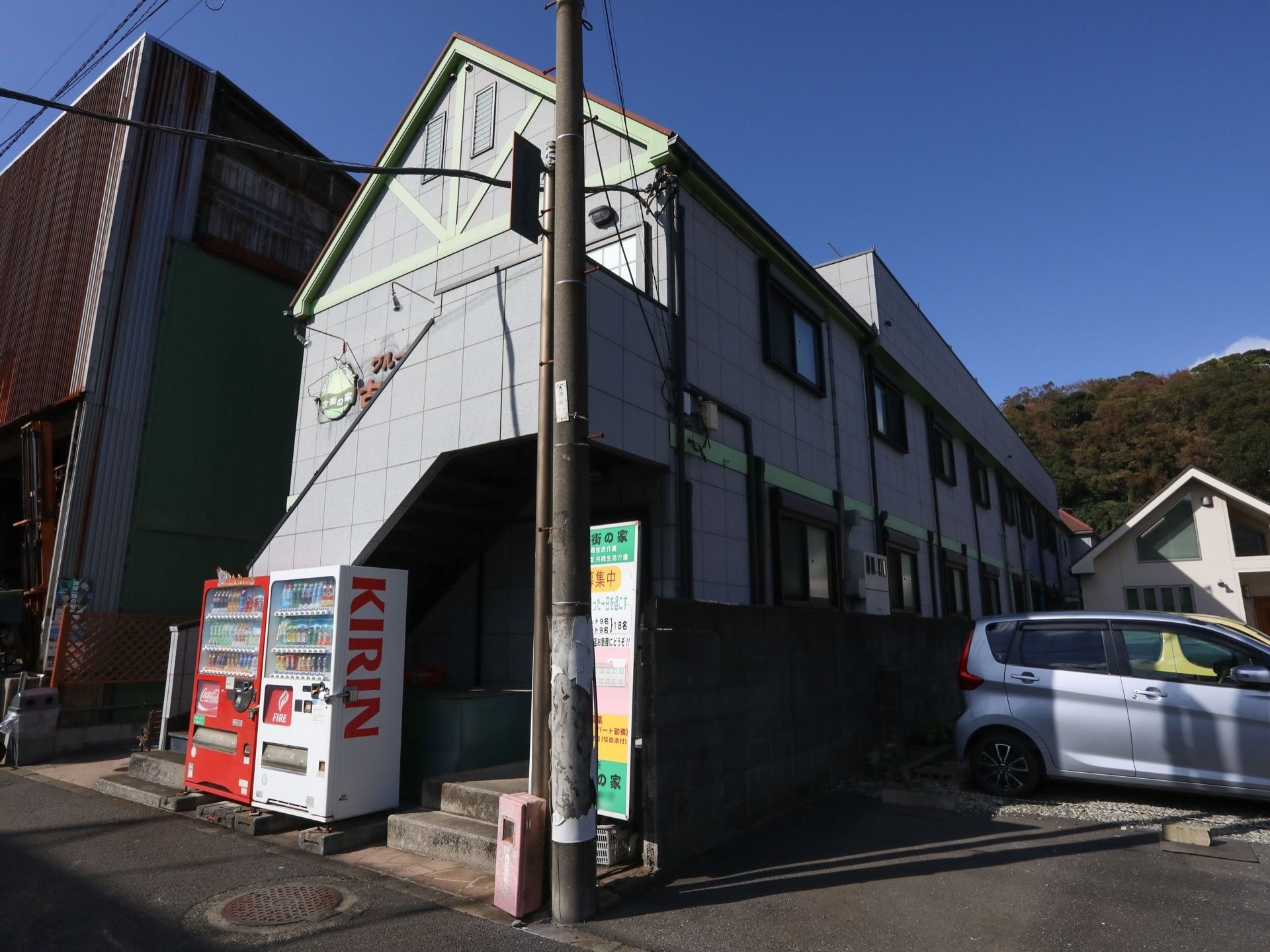 グレーの2階建ての施設の横に自動販売機が２台設置されている。また、道路に面したところに施設について書かれた看板が置かれている。