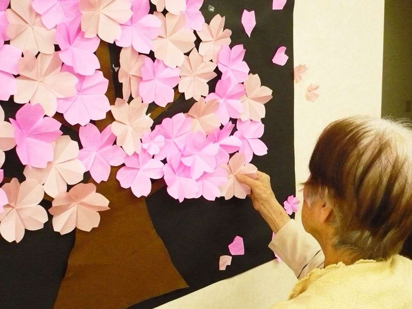 壁に貼られた大きな壁画。桜の大きな花が貼られていて、高齢女性が持っている花をその木に加えて貼ろうとしているところ。