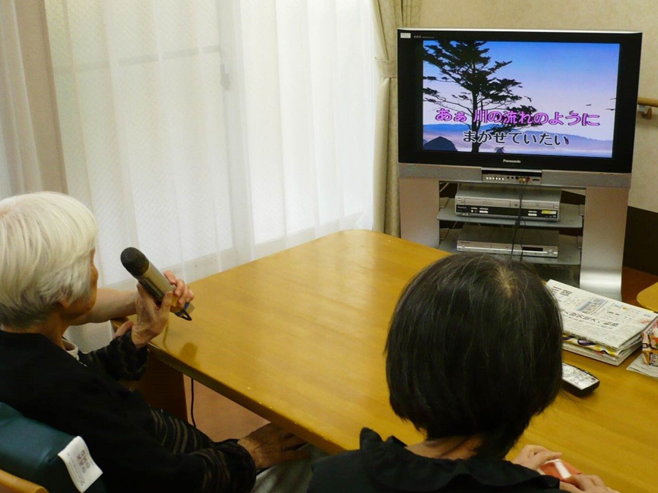 カラオケを楽しむ女性二人。左側には白髪の女性がいて、マイクを持ってテレビの方を見ている。その隣には黒髪の女性が座っている。