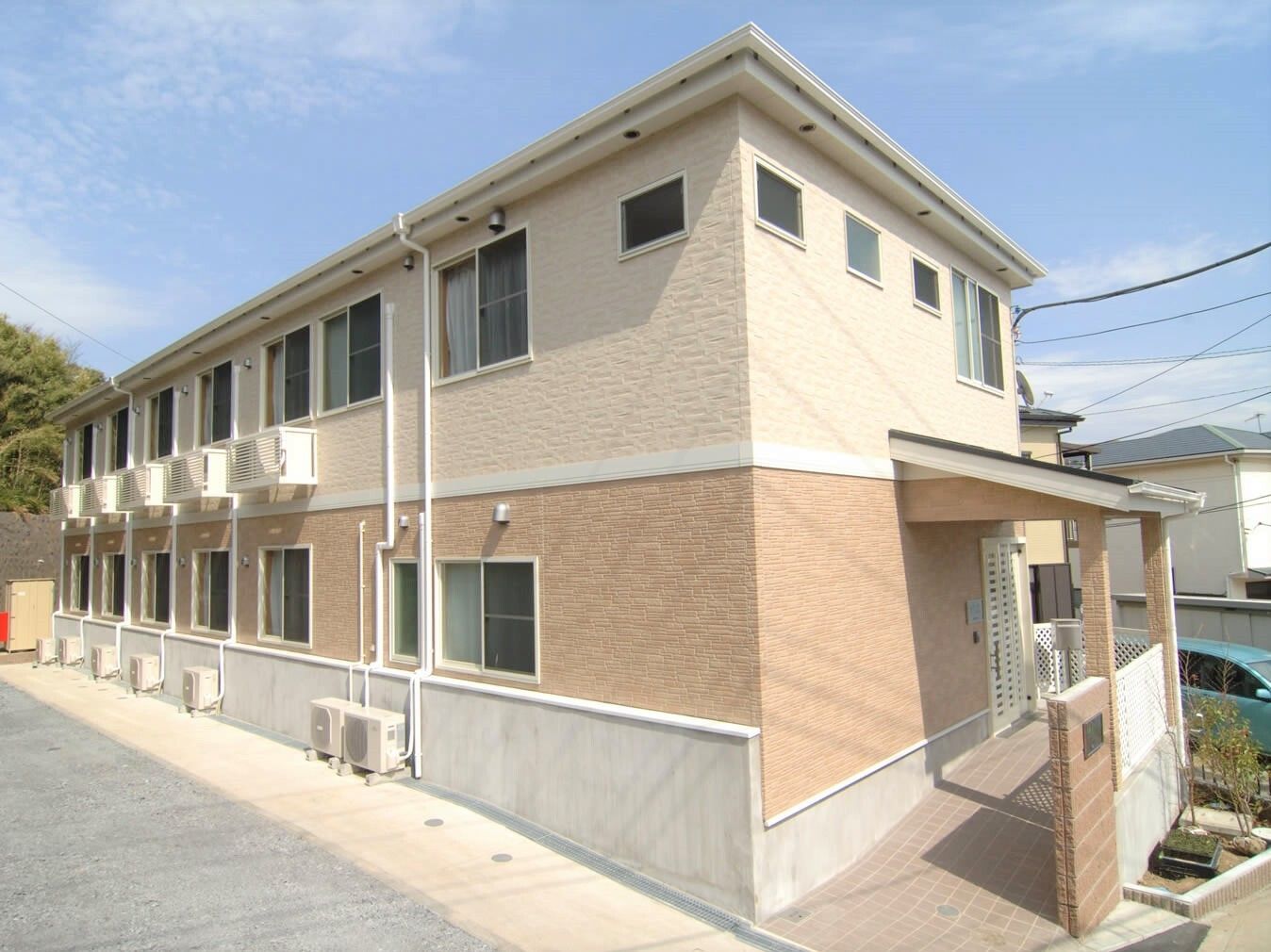2階建ての建物の周辺には緑が程よくあり、日当たりも良好。建物には屋根があり、玄関先にはスロープと屋根があり、奥には住宅も見える。