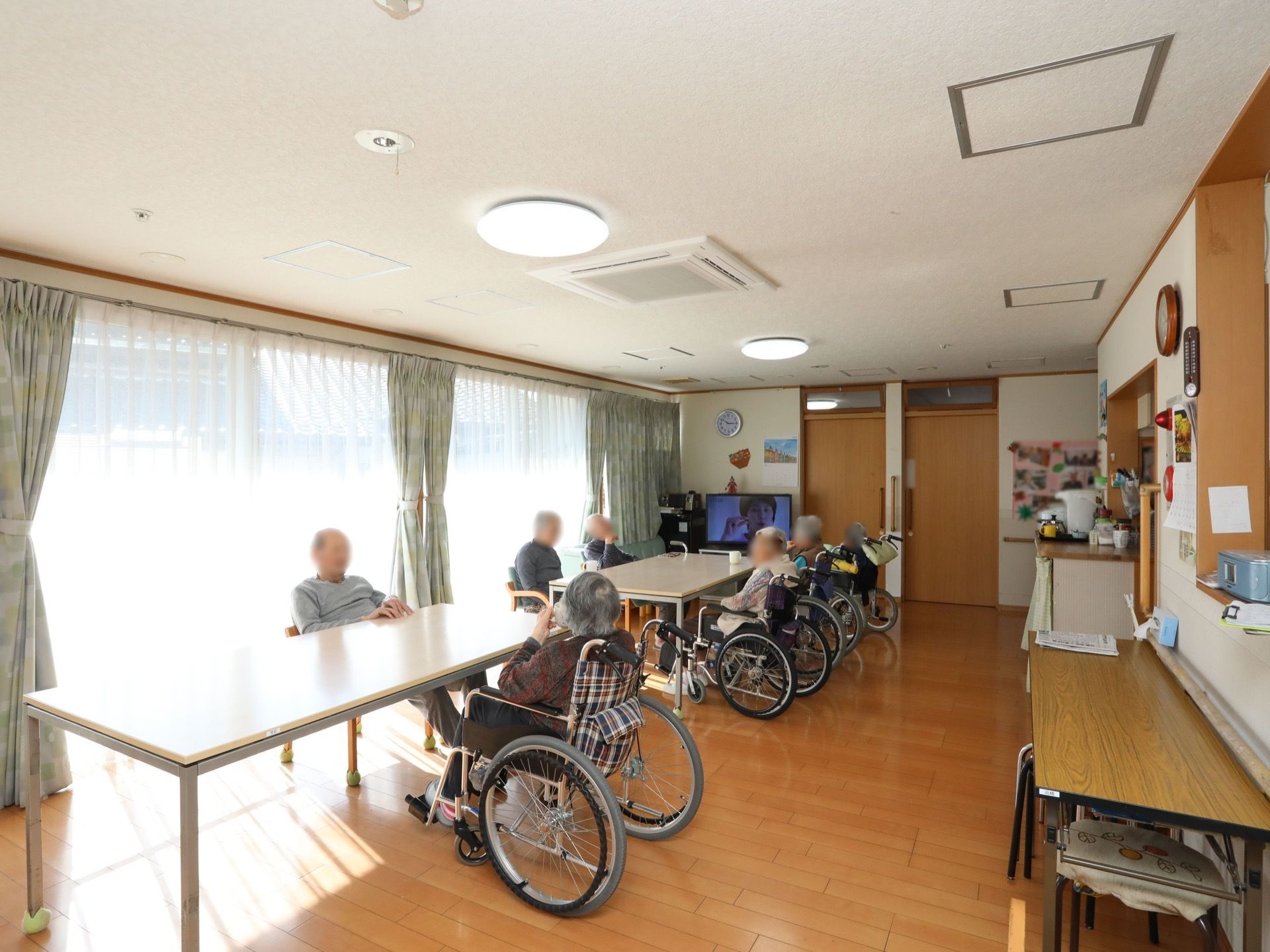 大きな窓にレースカーテンと遮光カーテンがつけられている。テーブルを囲んで入居者様が椅子や車椅子で座っている。