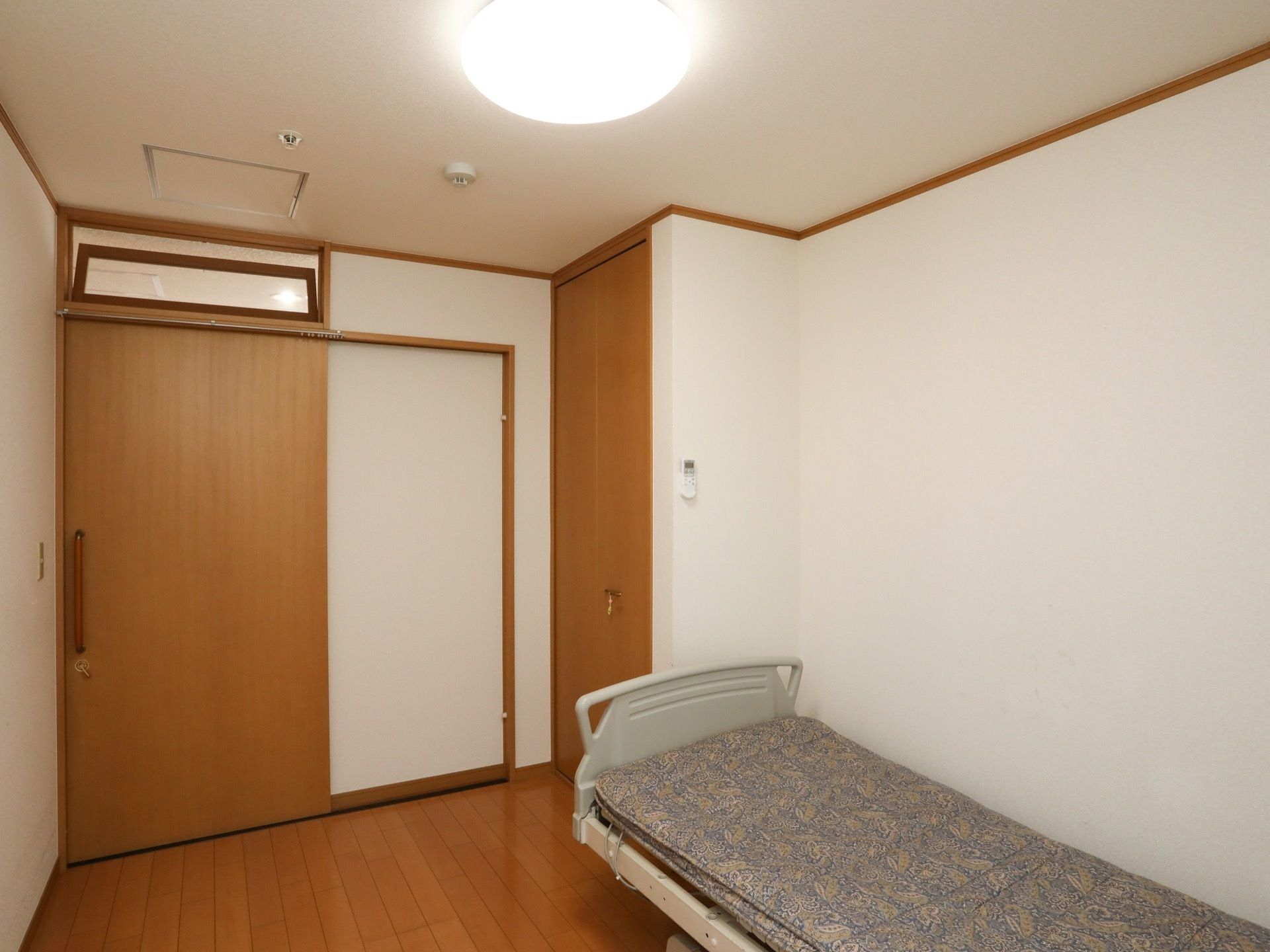 居室のドアは木目調で、スライド式である。床はフローリングで壁紙は白く、清潔感のある空間となっている。介護用ベッドがある。