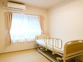レースカーテンとカーテンが付いたおおきな窓がある部屋には介護ベッドやエアコンがある。