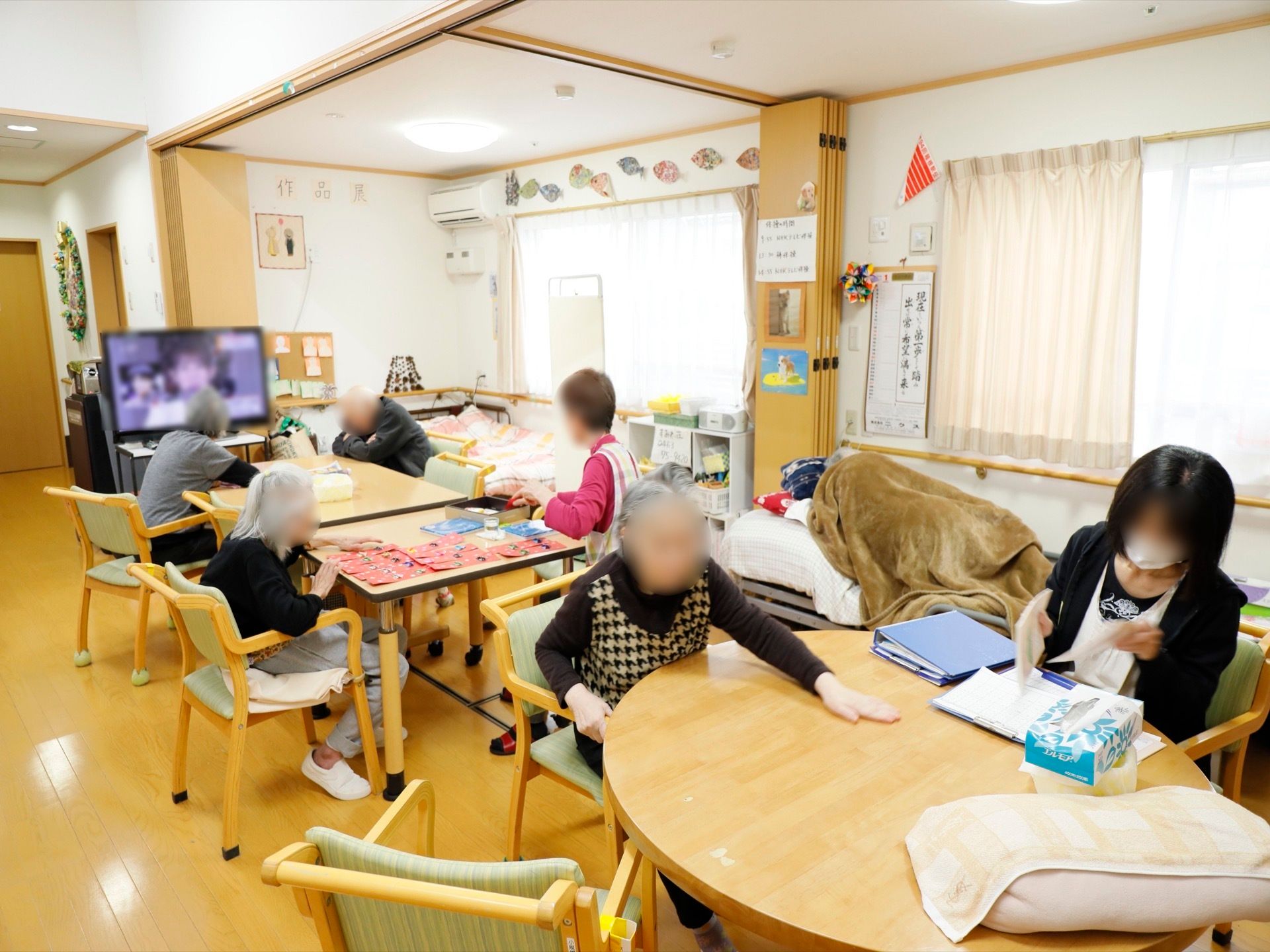 入居者様とスタッフが一緒にテレビを見ながら、作業をしている様子である。壁際にはベッドが設置されており、寝ている入居者様がいる。