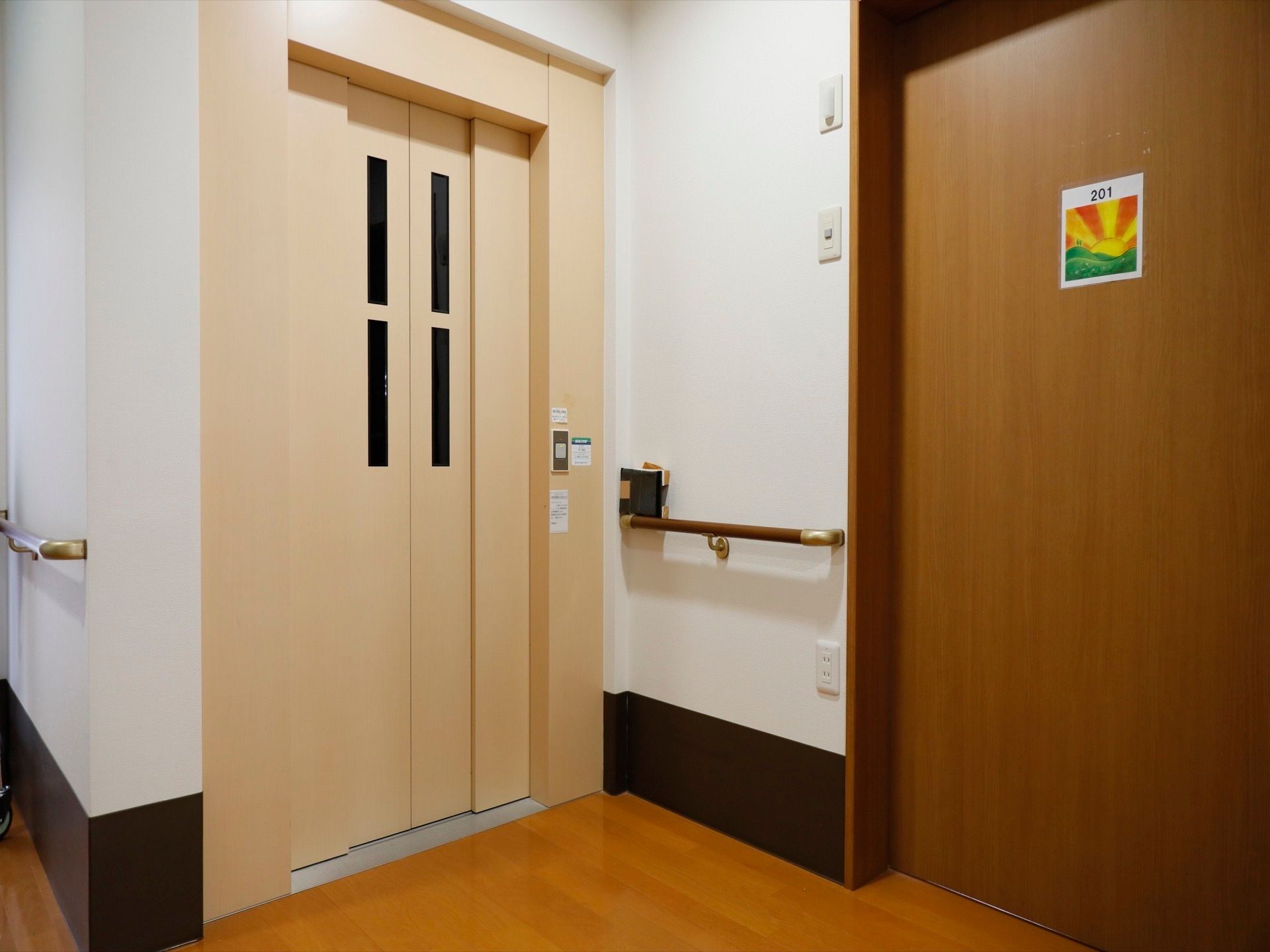 白とグレーのツートンカラーの壁とベージュの扉のエレベーターである。壁には手すりが取りつけられている。