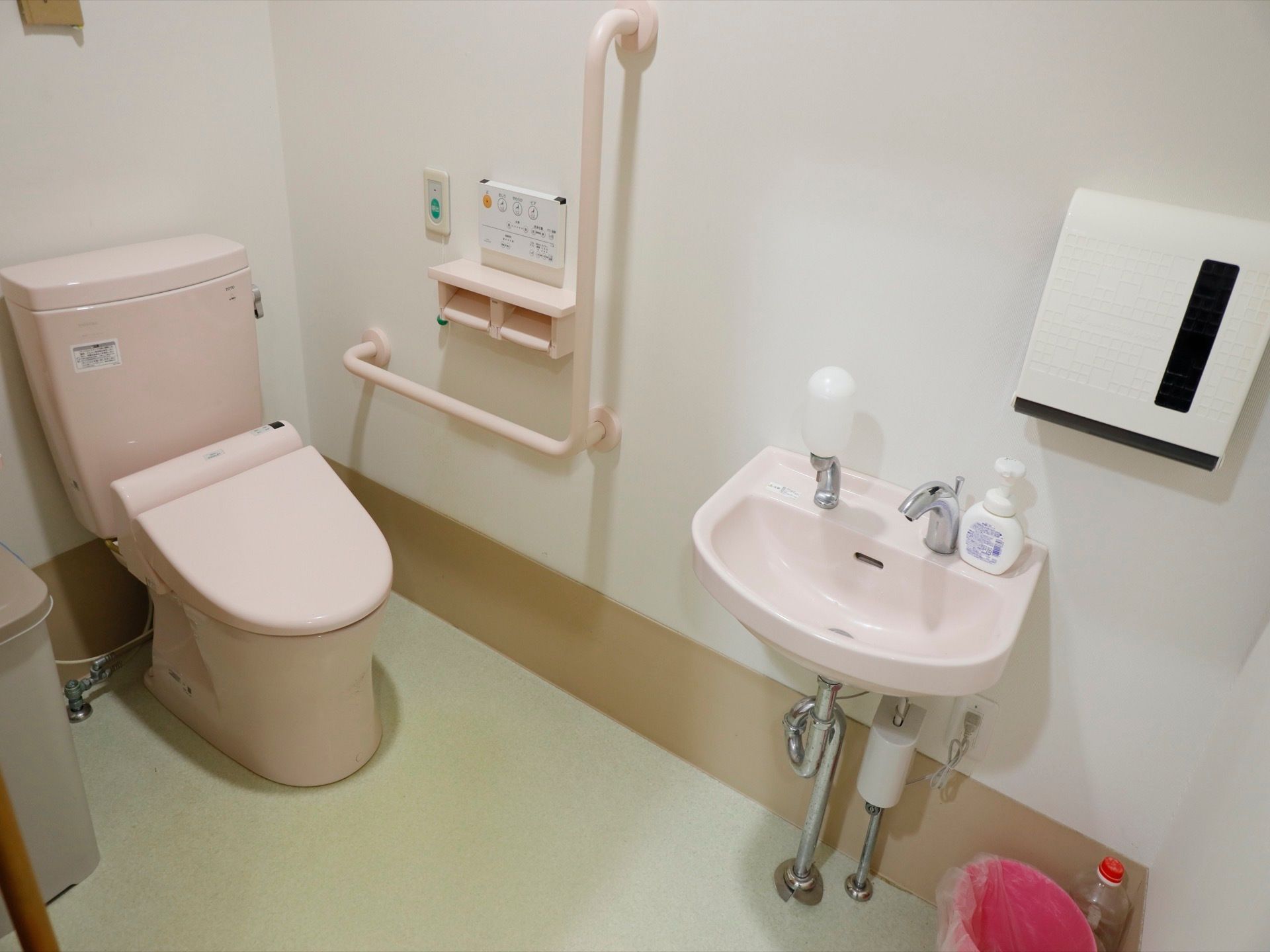タンク付きの洋式トイレである。壁には手すりが取りつけられており、手洗い場も設置されている。床にはごみ箱が置かれている。