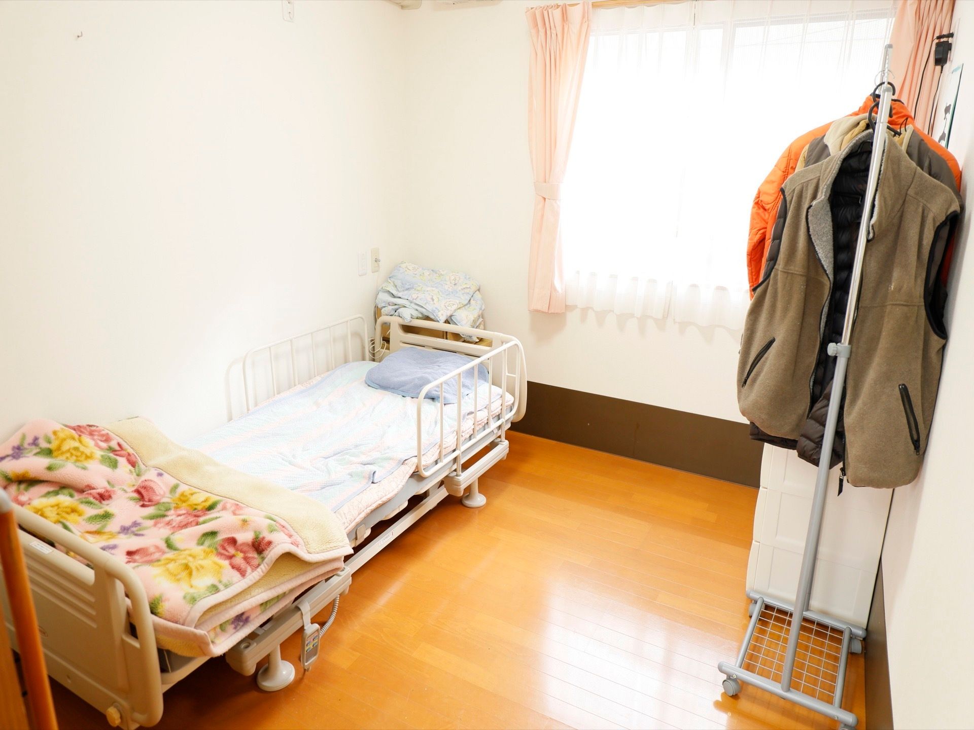 白い壁の居室に、白いフレームのベッドが置かれており、ベッドガードが設置されている。窓にかけられているカーテンは、優しいピンク色である。