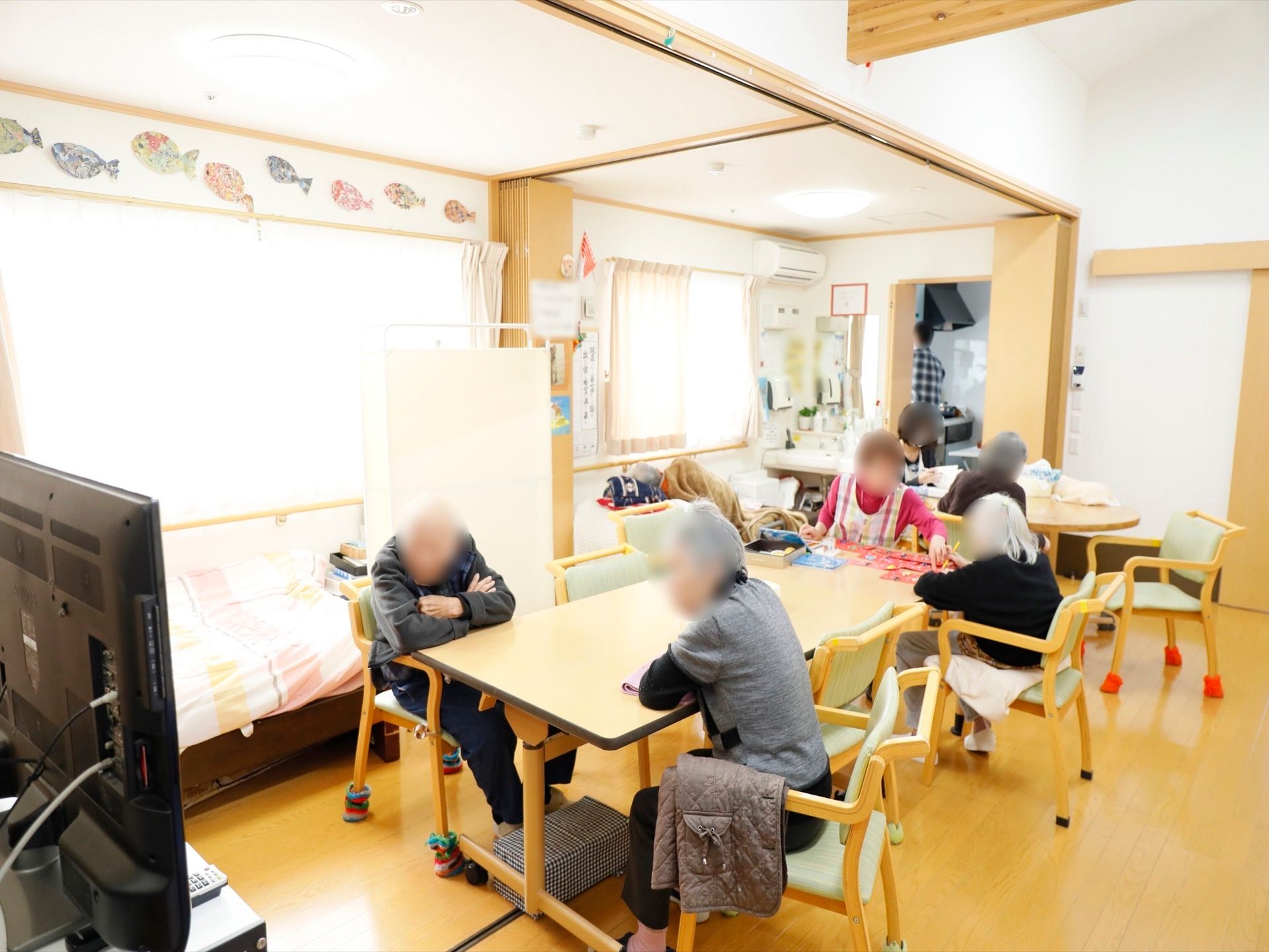 テーブルを囲むように入居者様とスタッフが座っている様子である。テレビが設置されており、壁には手作りの飾りが飾られている。