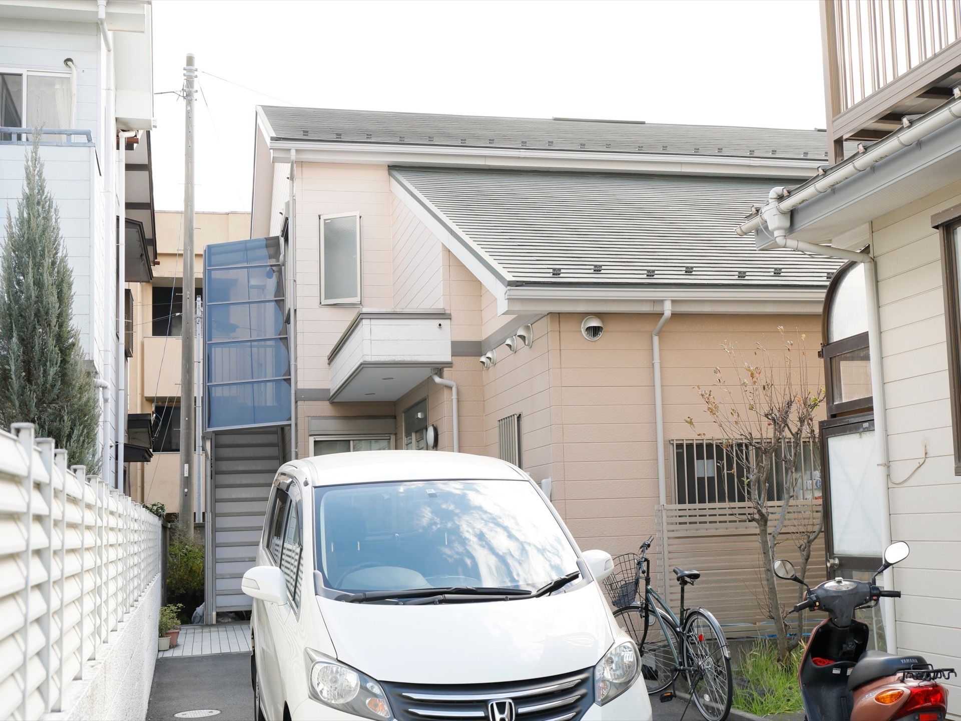 手前に車が１台停まっており、その奥に外階段の２階建ての建物がある。車の横には、自転車と原付バイクが停められている。