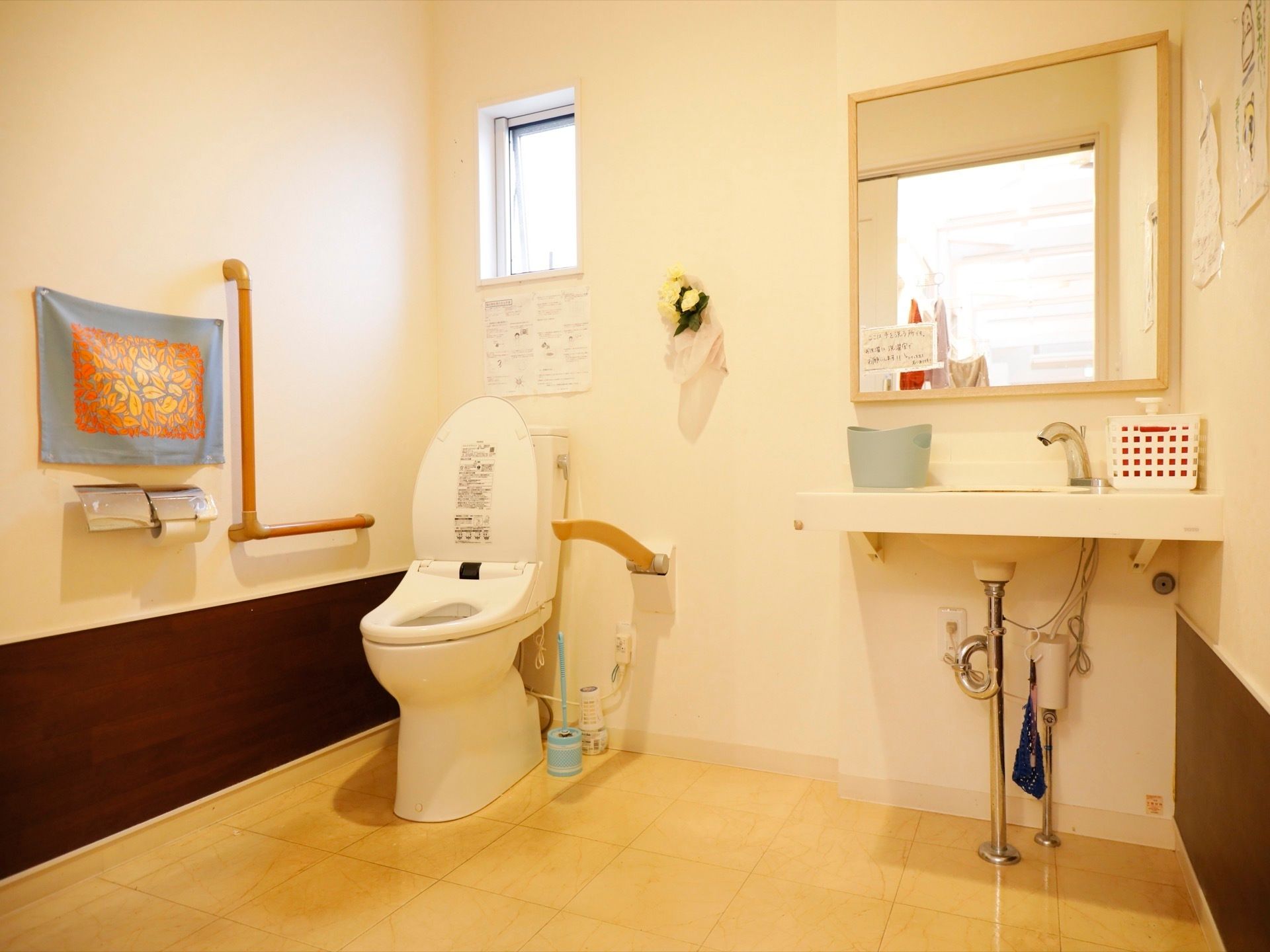 洗面台が備えつけられた清潔感のあるトイレである。上部には小窓が設置されており、便器の両サイドには手すりがある。