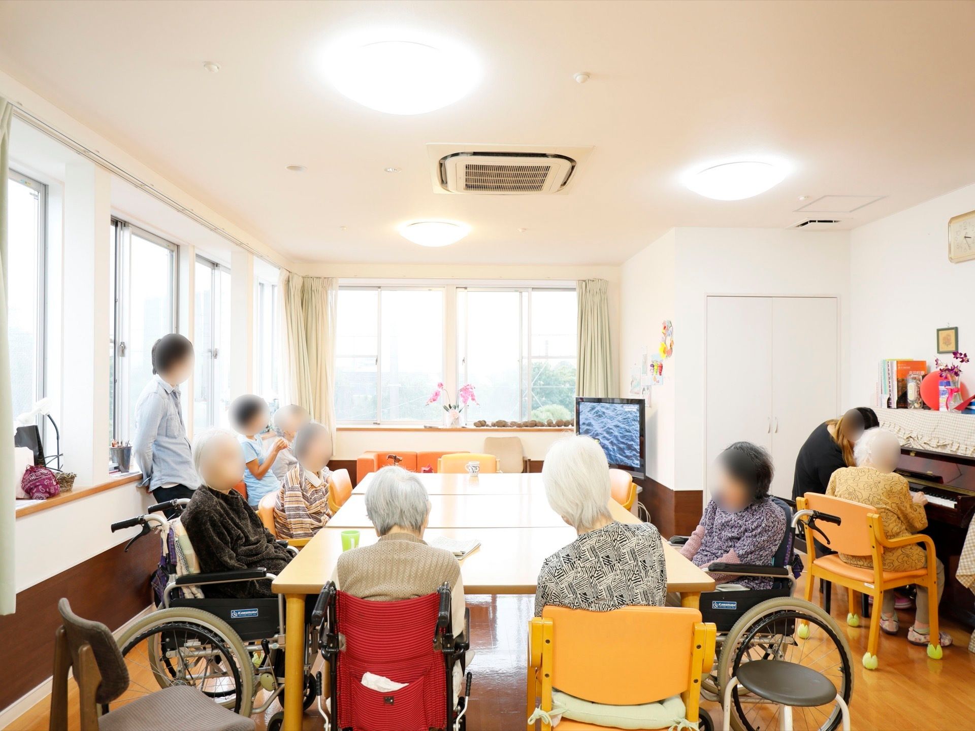 大きな窓のある日当たりの良いリビングスペースで、フロア隅にテレビがある。食卓を囲んで入居者様がくつろいでいる。