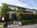 道路とは植樹で隔てられた、２階建ての建物である。玄関へ向かう階段には手すりが設置され、階段の横には、駐輪場が設けられている。