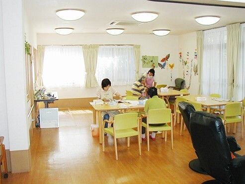 ライトグリーンの椅子がセットされた木製のテーブルが複数置かれ、スタッフと入居者が思い思いの場所にいる。
