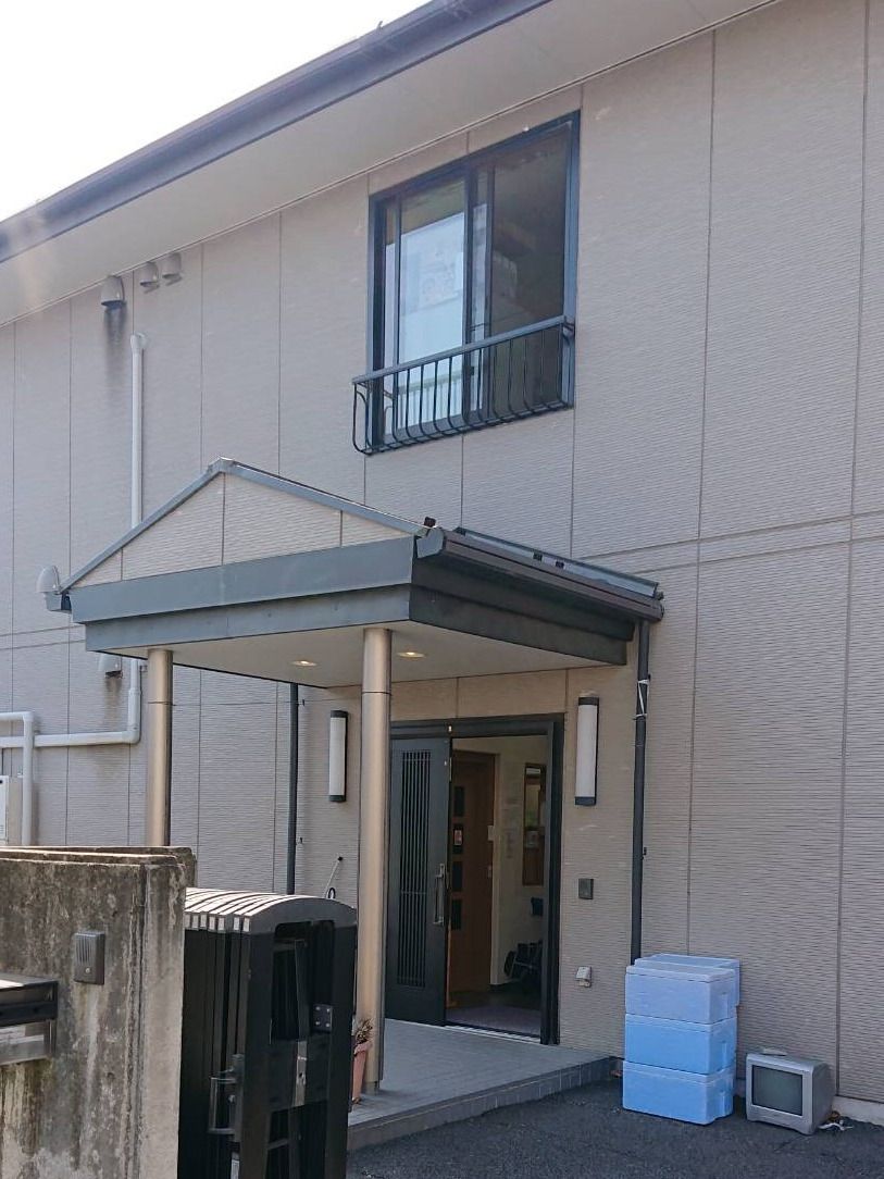 グレー系の外観の２階建ての建物は玄関に屋根があり、草木が周りに植えられている。