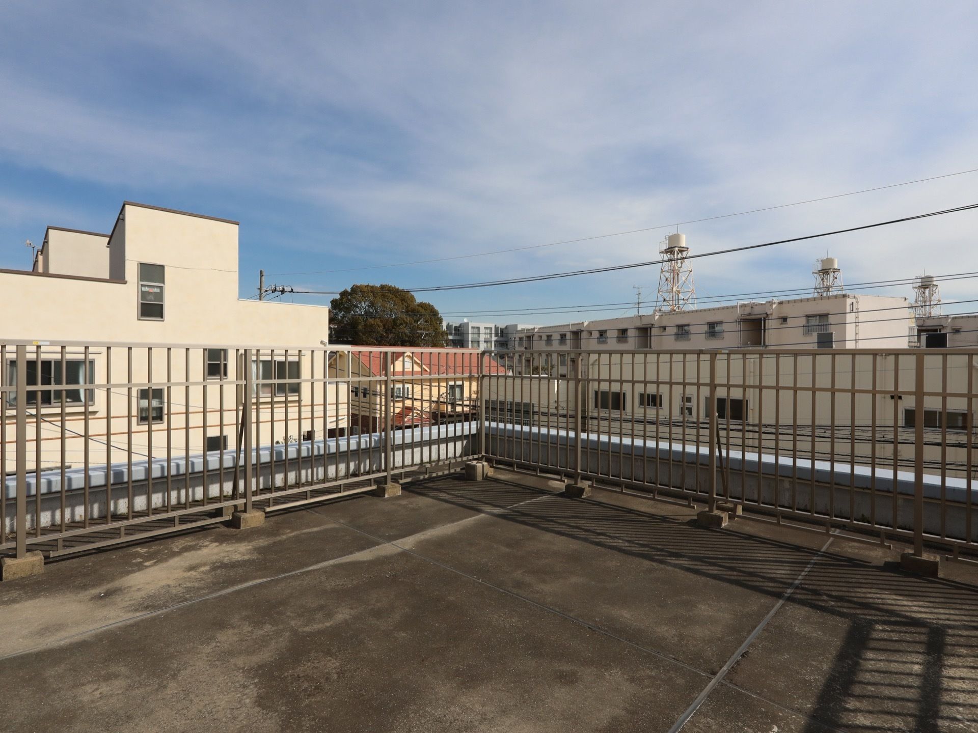 屋上の周囲に背の高いフェンスが設置されている。近くの集合住宅や背の高い木が見える。空には薄い雲が出ている。