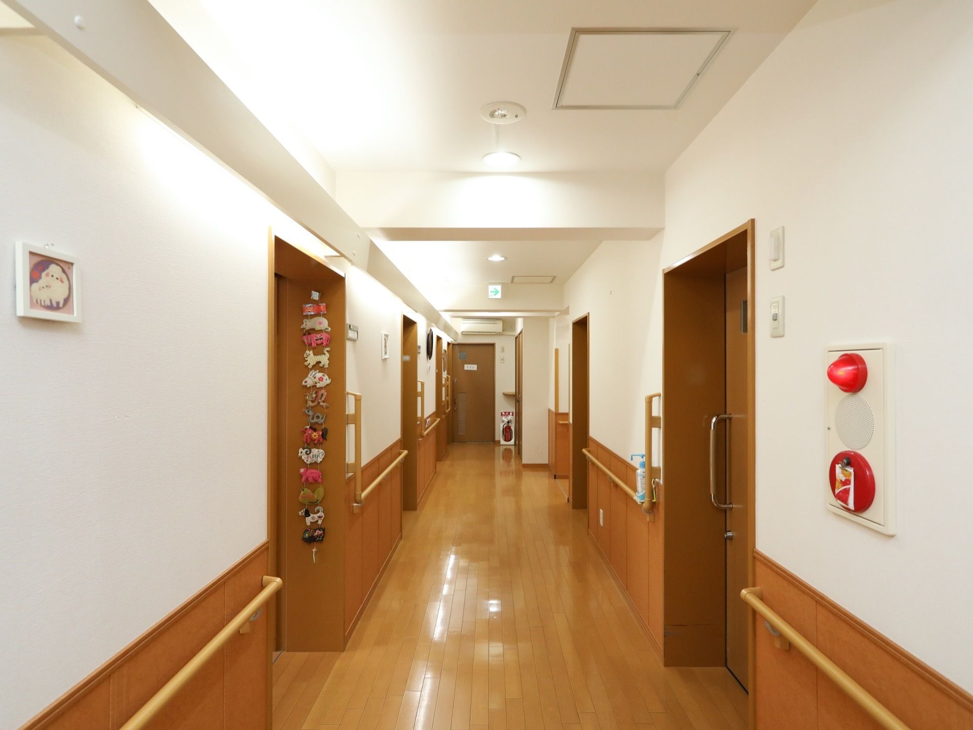 壁や天井の白色と、床面や居室入口扉の茶色のツートンカラーの廊下である。天井の片側は間接照明になっている。