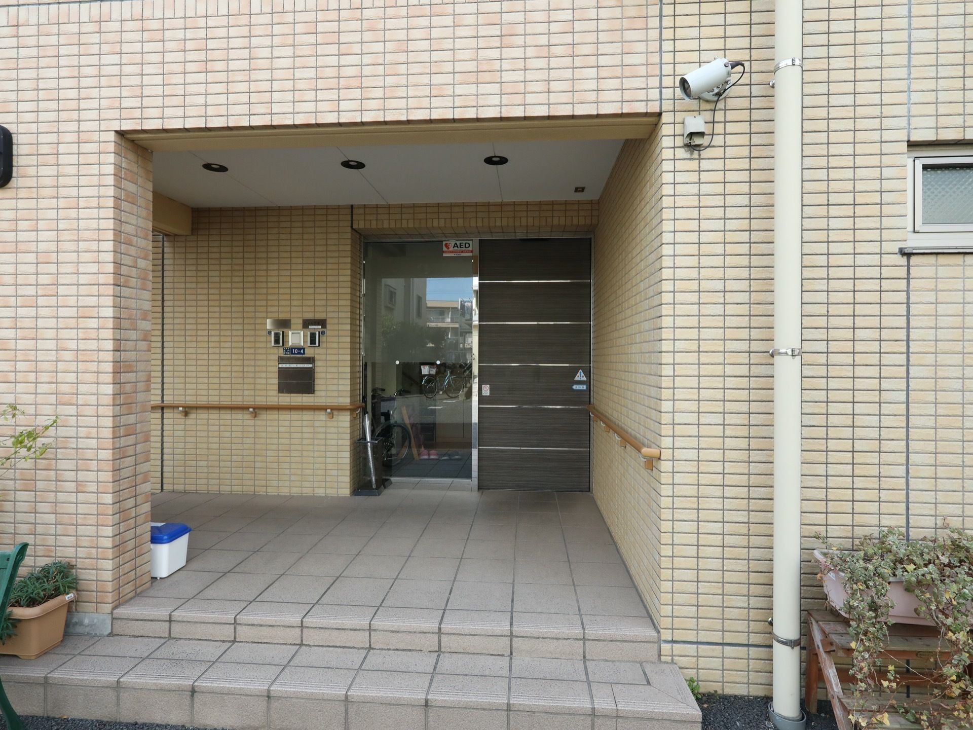 玄関前の壁の上の方に防犯カメラが取り付けられている。玄関扉の横のガラスにAED設置のステッカーが貼られている。