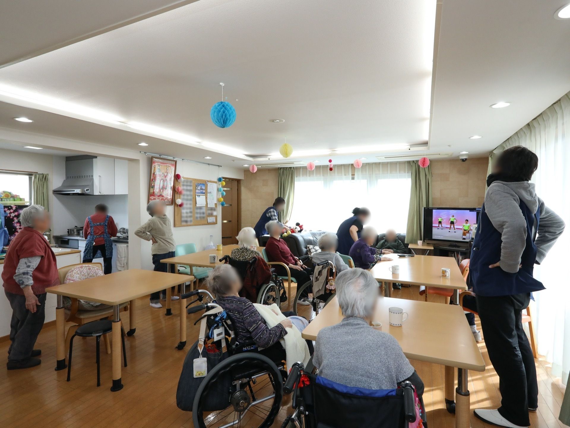 食堂に多くの入居者様が集まっている。窓際に立っているスタッフと一緒に、テレビ番組の体操を行っている。