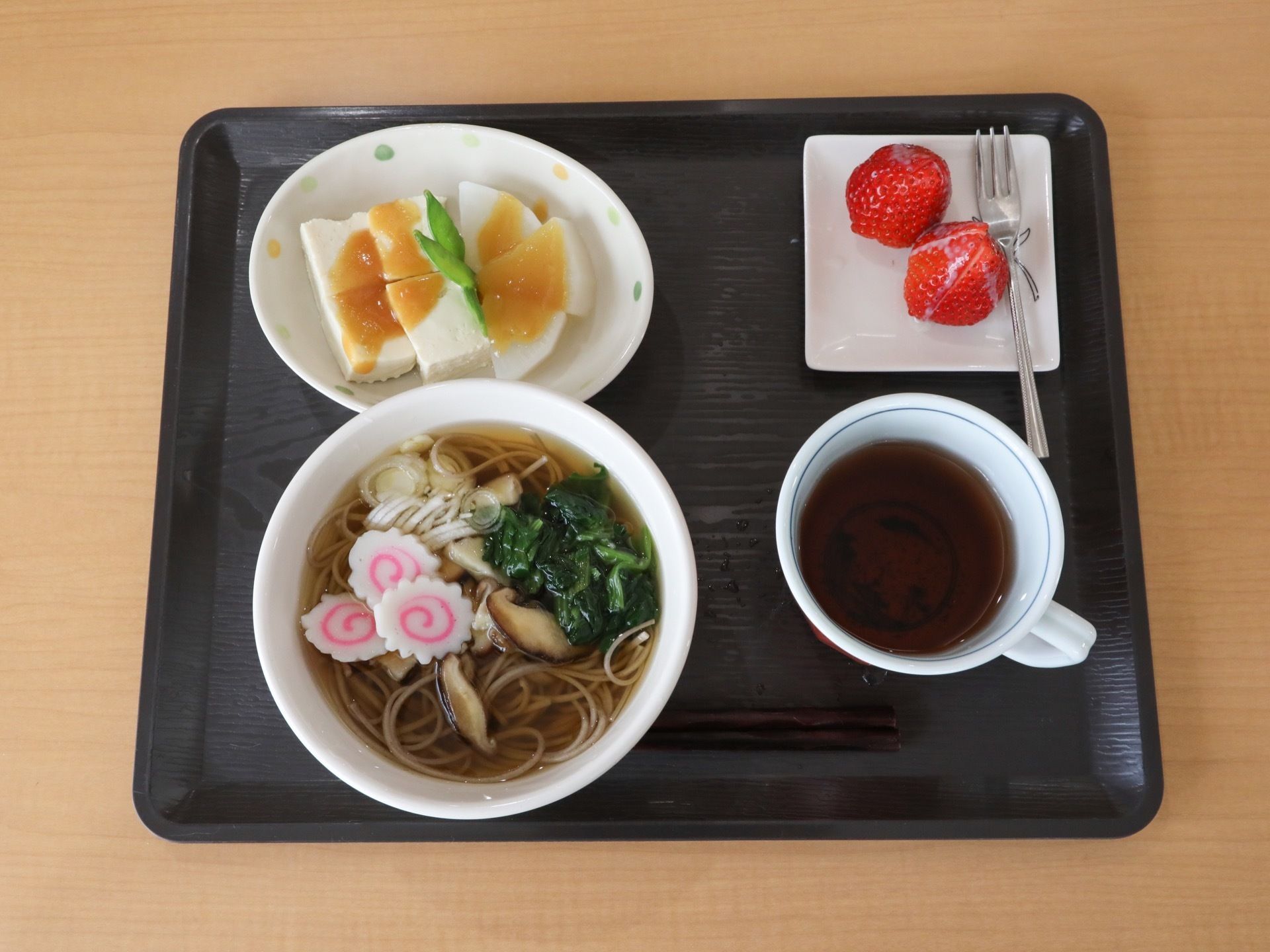 長方形の黒色のお盆に、そば、豆腐、いちごのお皿が載っている。手前に、飲み物が入ったカップが置かれている。