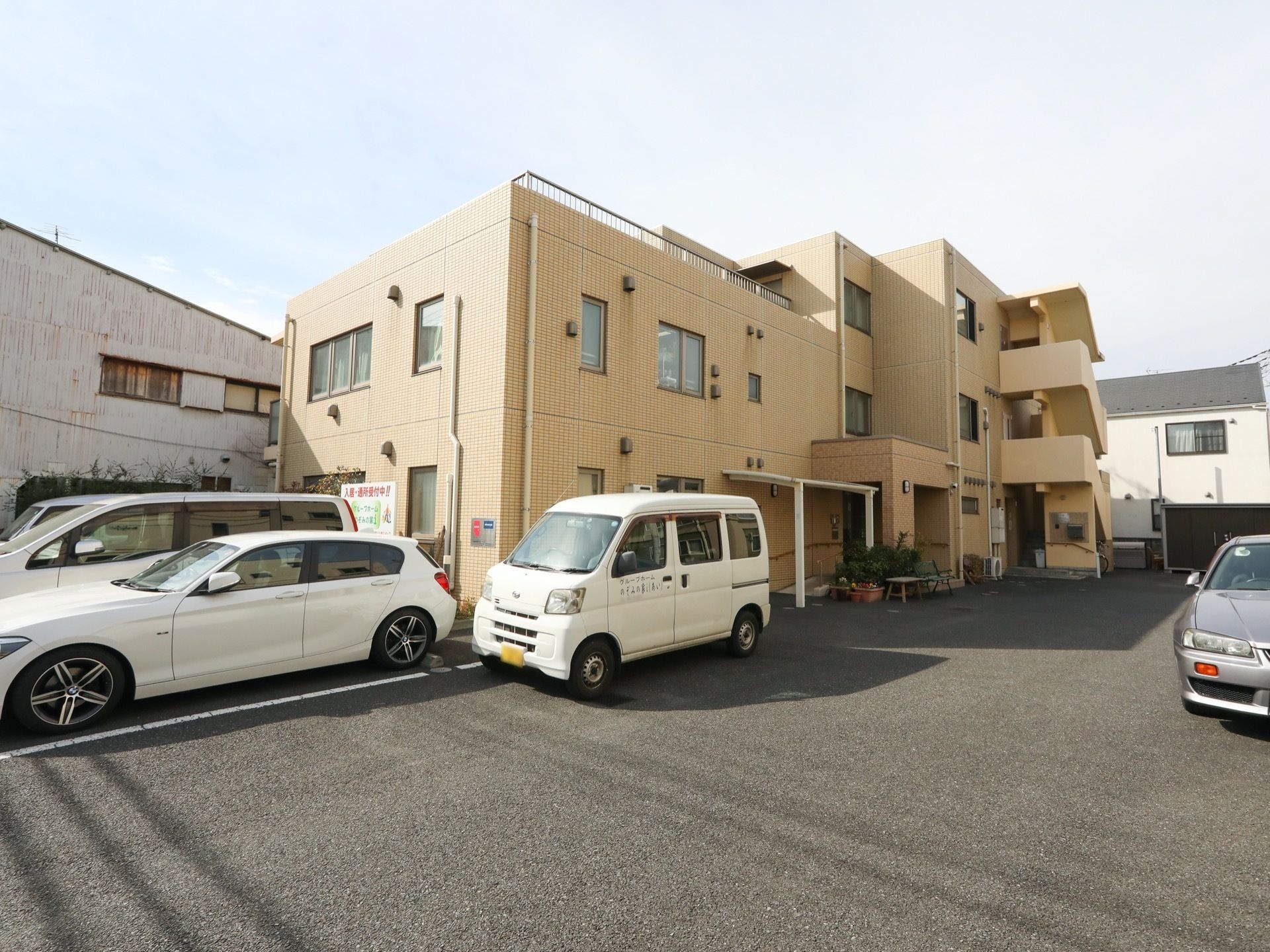 黄系統の色の外壁を持つ、一部３階建ての施設である。敷地内に駐車スペースがあり、車が何台も停まっている。