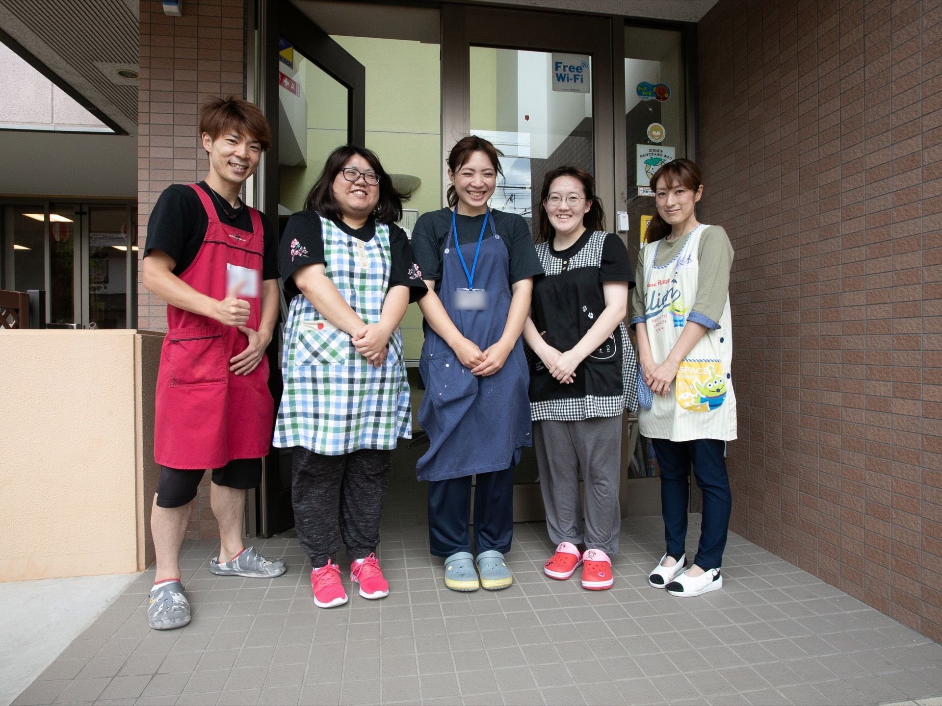 施設の玄関前で女性が４名、男性が1名の計５名のスタッフが笑顔で並んでいる。全員エプロンを着用している。