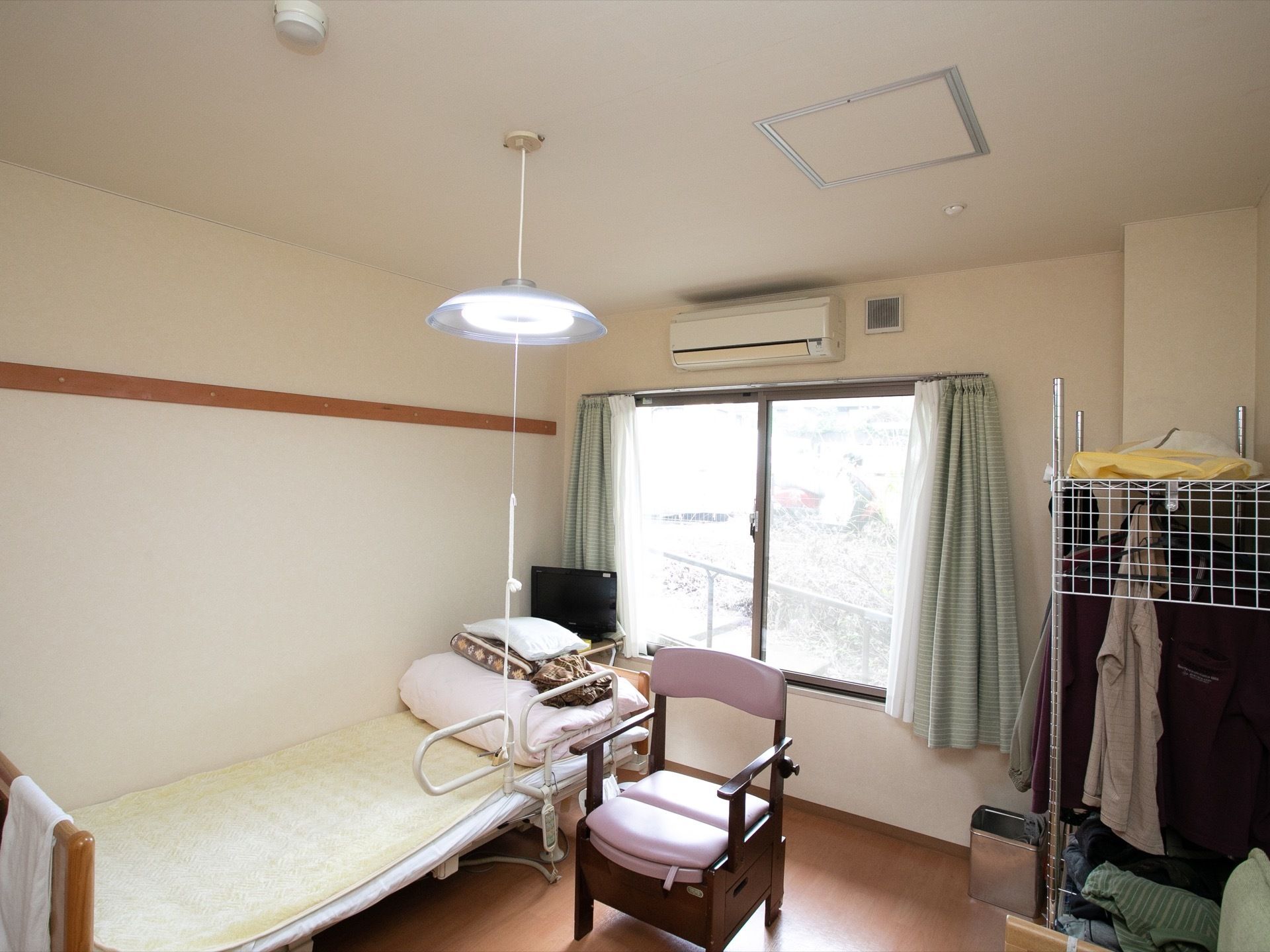 ワンルームタイプの居室である。介護用ベッドとポータブルトイレの他、壁にはエアコンが設置されている。ハンガーラックが持ち込まれている。