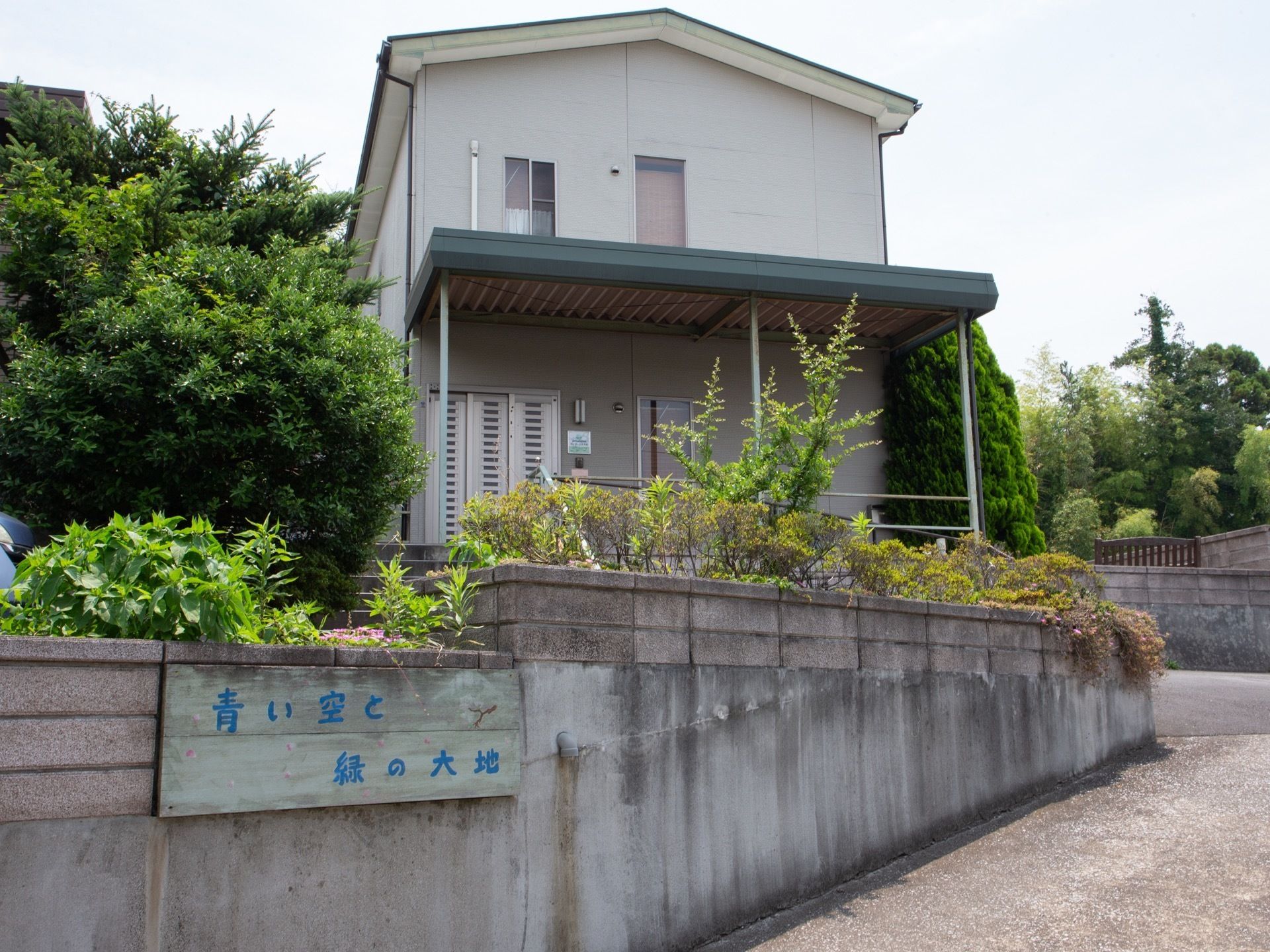 ゆるやかな上り勾配の先に、一軒家のような２階建ての施設が建っている。手前の塀に、木製の施設看板が設置されている。
