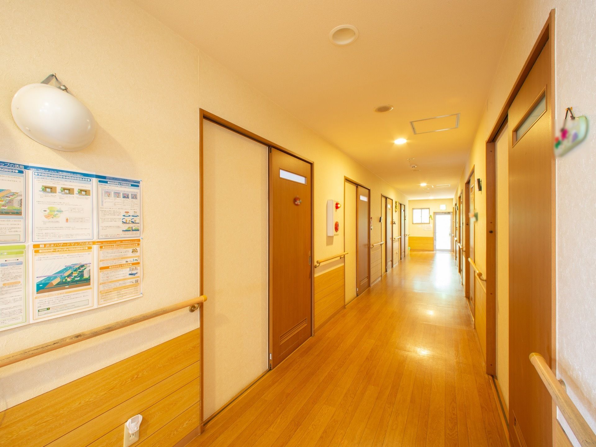 壁に緊急災害通報装置や手すりが設置されている居室前の廊下である。清掃が行き届き、すっきりと見通しが良い。