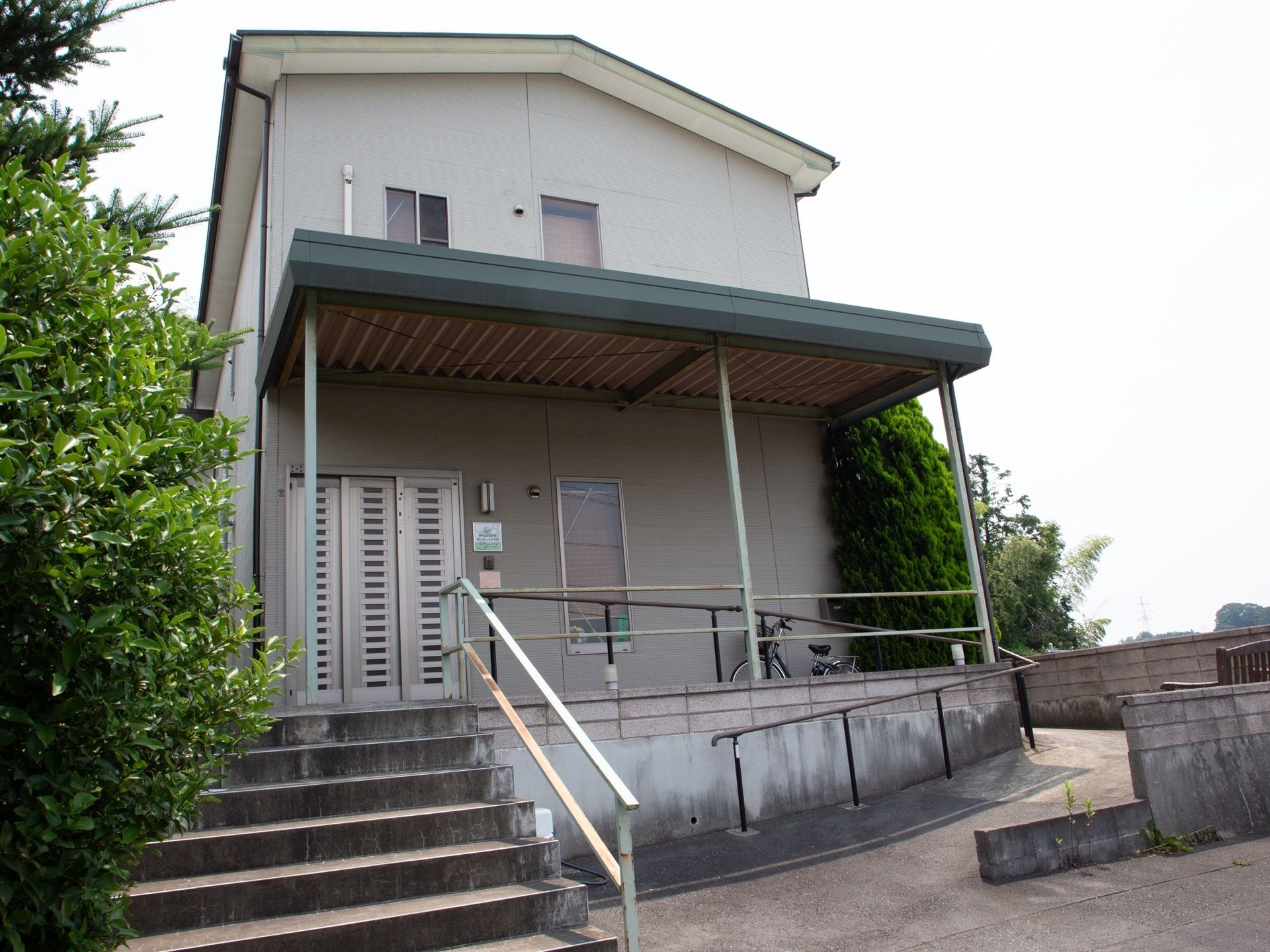 ゆるやかな勾配を上り進むと、一軒家のような佇まいの施設がある。３枚引き戸の玄関が設置されている。玄関前には大きな屋根が取り付けられている。
