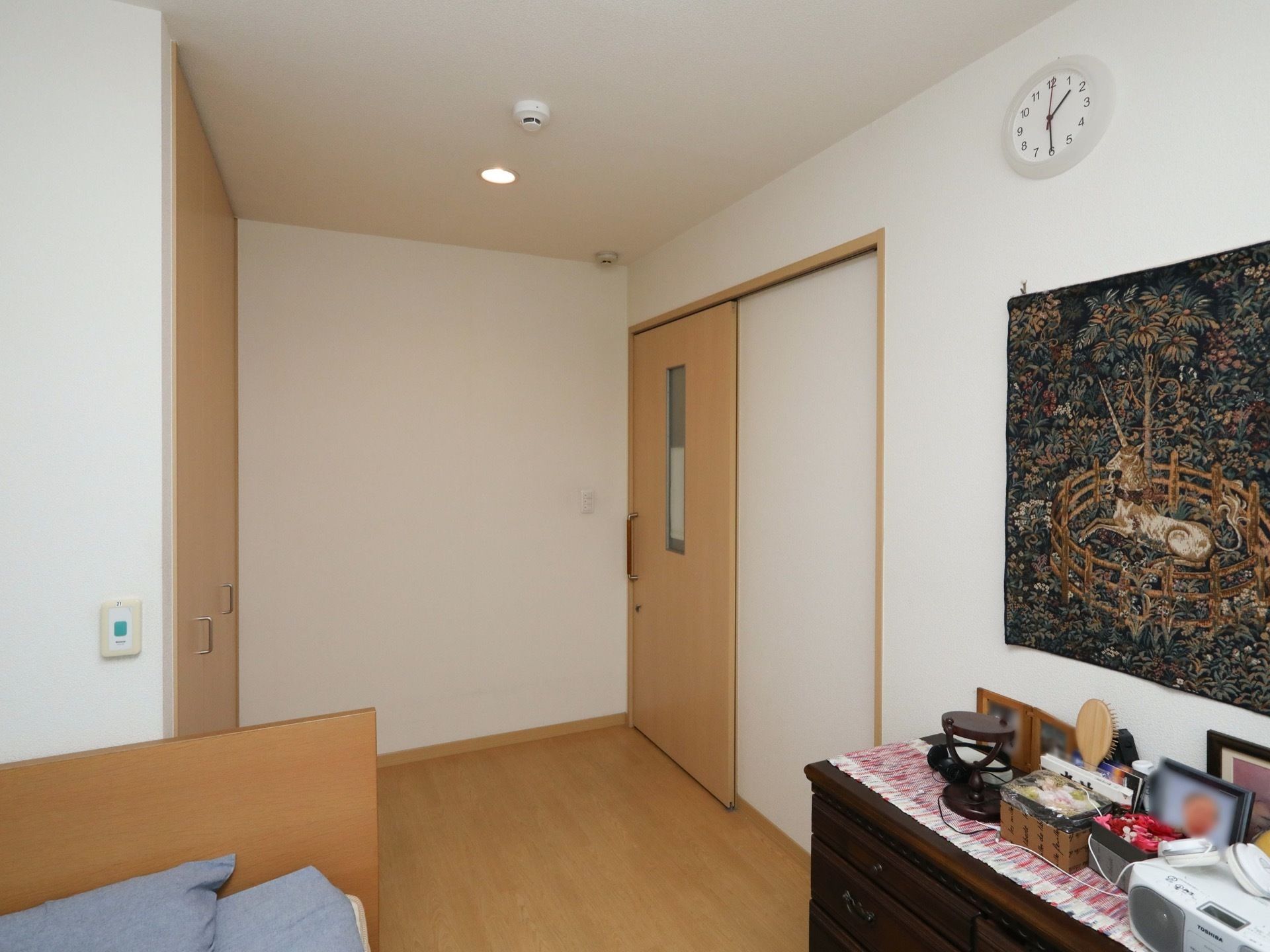 居室の入口はスライドドアである。壁には絵が飾られており、その前の棚の上にはラジカセや写真などが置かれている。