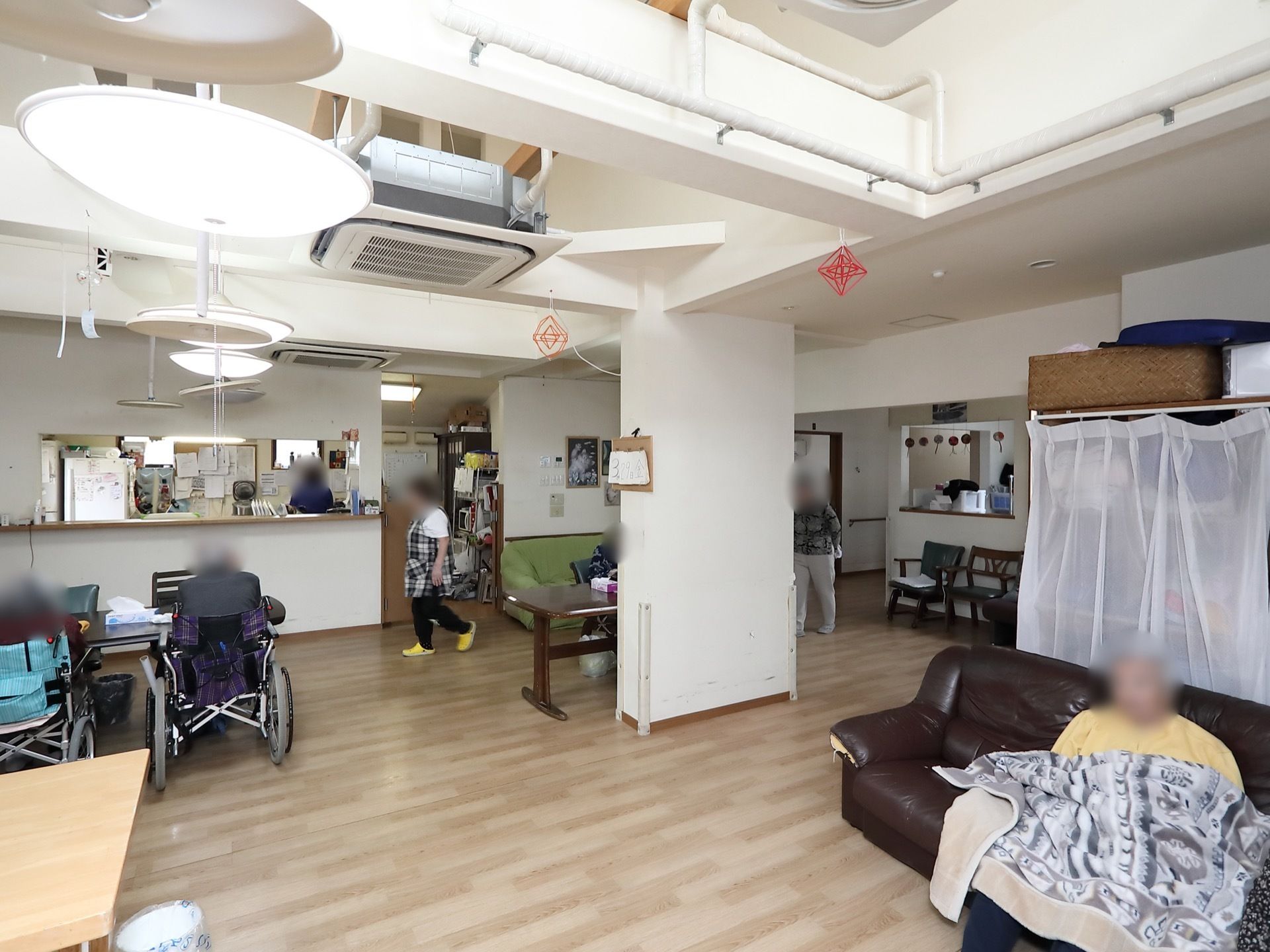 右手前に黒いソファが置かれ、入居者様がくつろいでいる。奥にはスタッフが常駐する事務所があり、スタッフが行き来している。