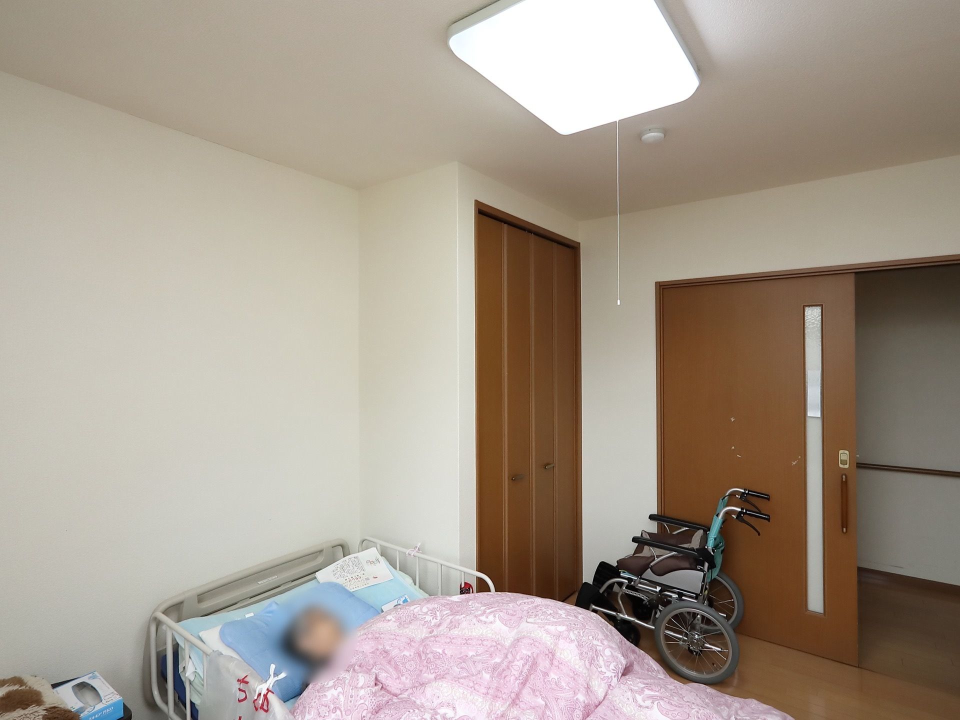居室の入口は引き戸になっており、入口前に車椅子が置かれている。折れ戸式の収納が左手にあり、その前にベッドが置かれている。