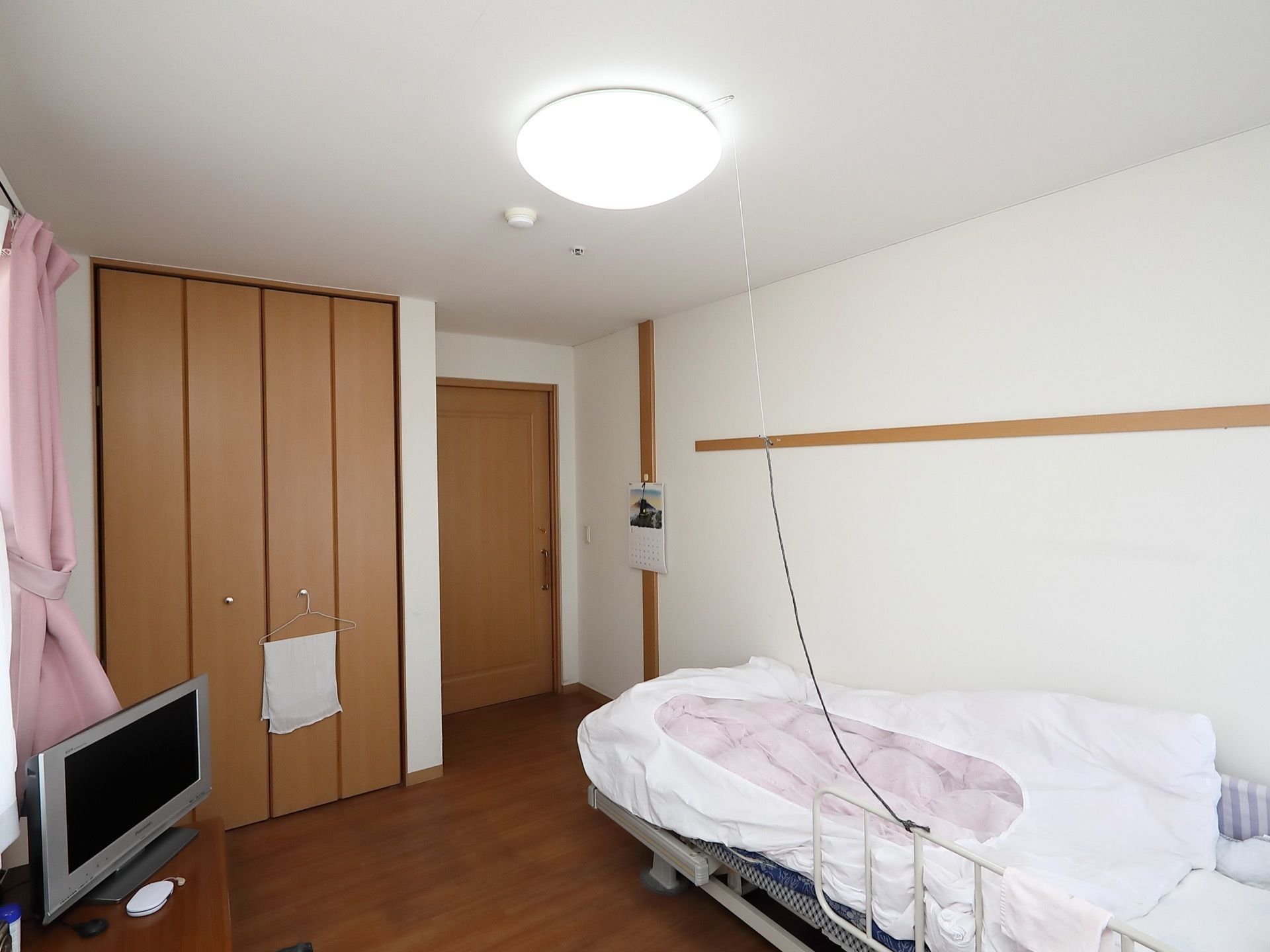 テレビが設置されている居室で、壁にカレンダーが掛けられている。天井には紐付きの照明が取りつけられている。
