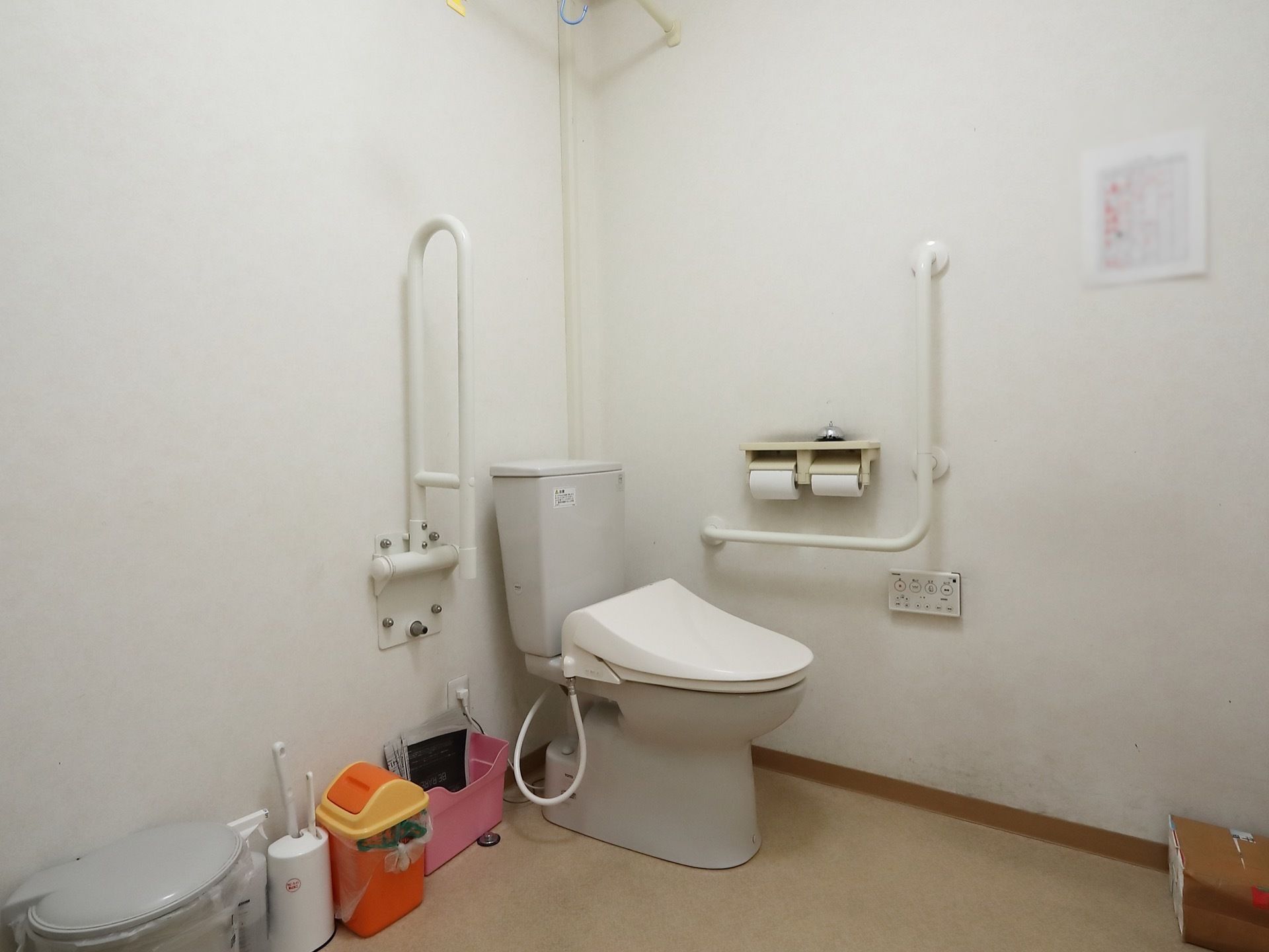 白を基調とした清潔感のある個室トイレである。トイレットペーパーホルダーの上には、ベルが置かれている。