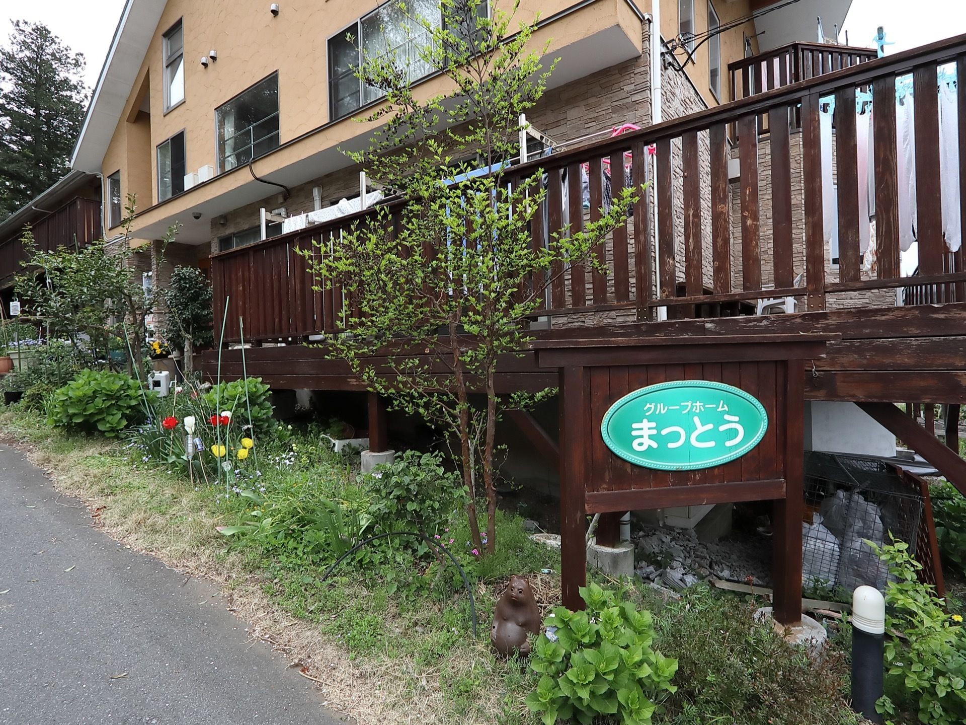 建物とアスファルトとの間に、洋風の庭木や草花が植えてあり、施設名称を示す看板が設置されている。デッキに洗濯物が干してある。