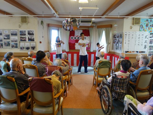 出演者が催し物を行っている様子とそれを眺める高齢者、家族、スタッフ