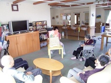 テレビが置かれた部屋で紙芝居を行う女性と、それを取り囲む高齢者たち