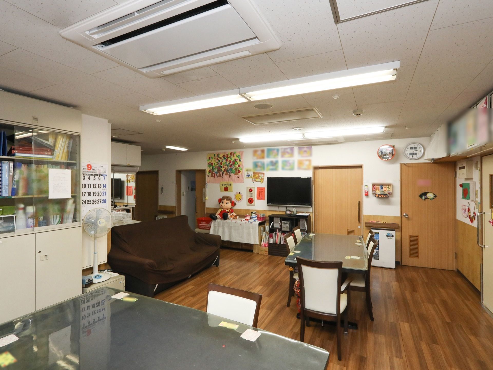 茶色の木目調床、白色の壁に造られた共有スペース。テーブル席を用意。壁側には、テレビが置かれている。