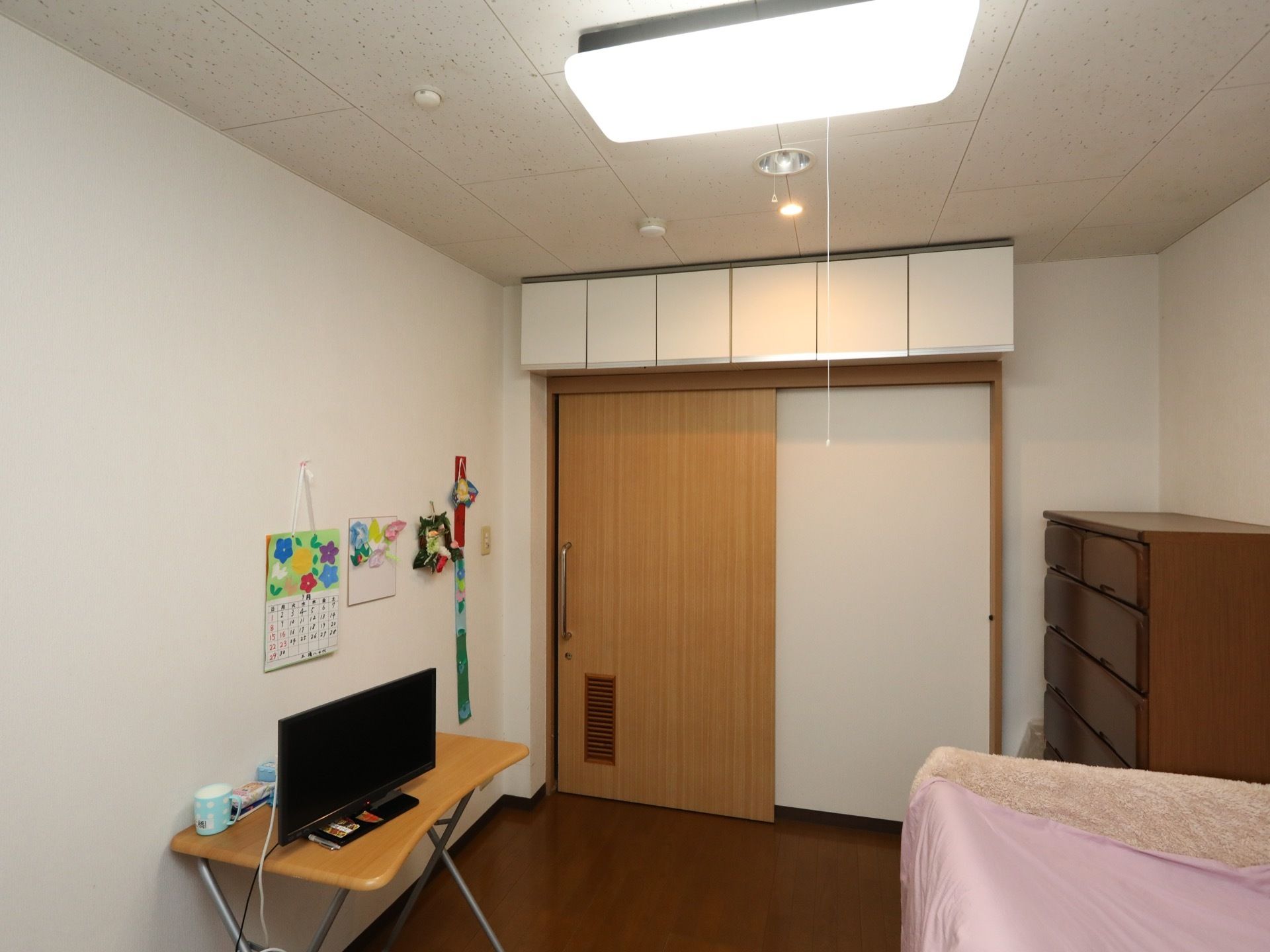 居室の扉上部に、収納スペースが備えられている。四角型の照明が設置されてある。デスクの上には、テレビも完備。