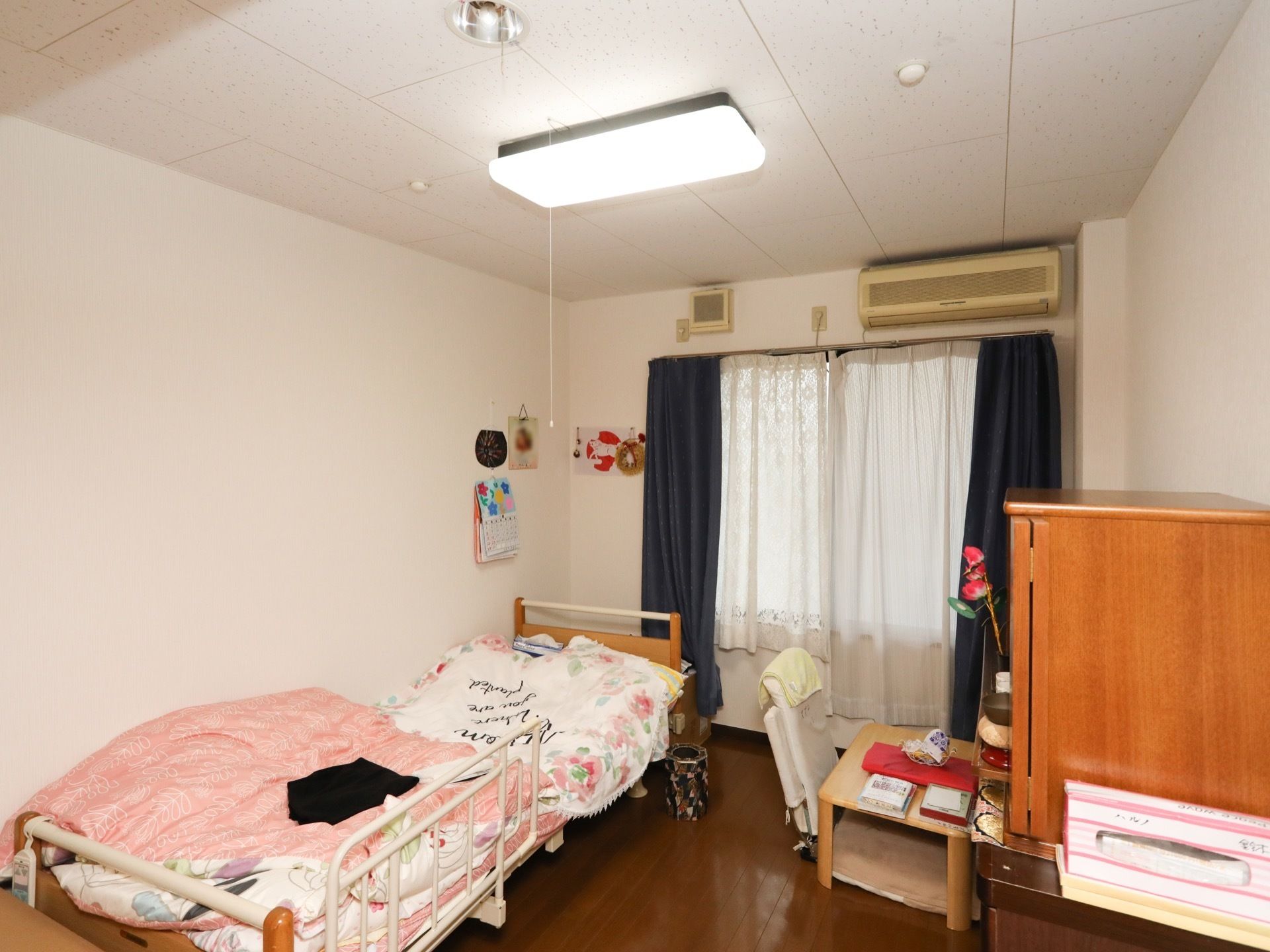 広々とした居室である。窓の上部に、エアコンが備えられている。サイドガードの付いたベッドも置かれている。