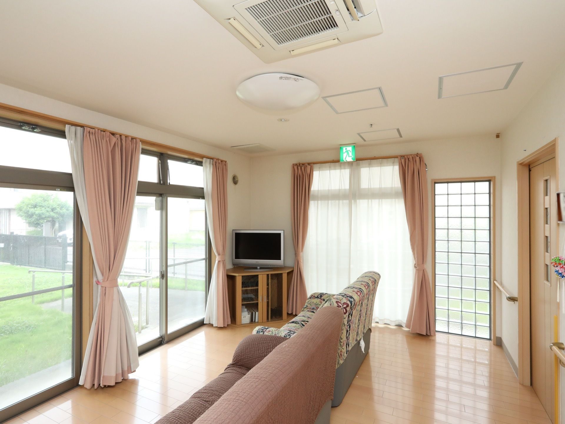 窓に淡いピンク、レースカーテンが備えられている。上部に非常口誘導灯がある。コーナーにはテレビが置かれている。